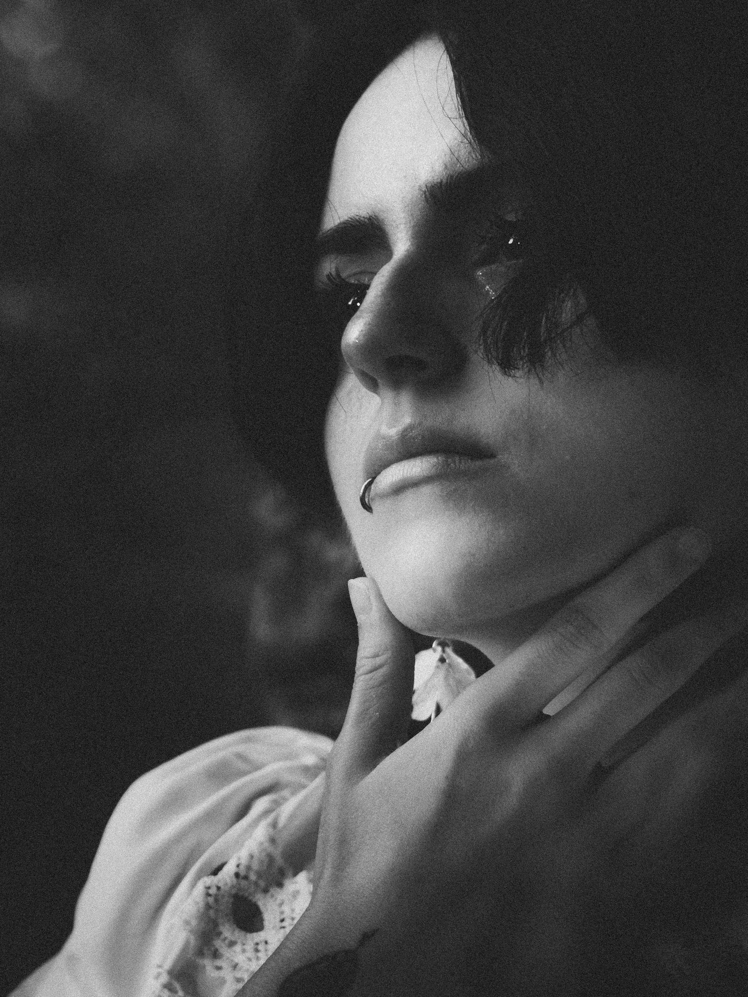 Black and white portrait of a person with dark makeup, gazing thoughtfully while touching their chin.