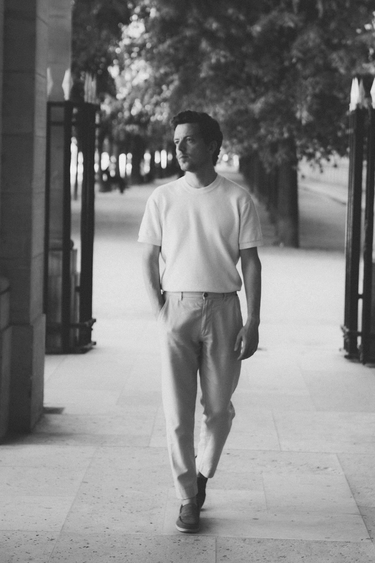 Black and white photo of a man walking confidently in a white t-shirt and trousers along a tree-lined pathway.