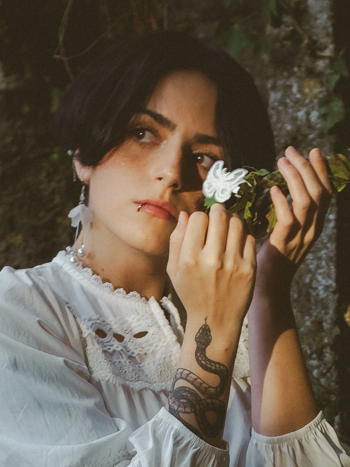 Young woman in white dress holds white flower, featuring snake tattoo on hand, posing outdoors near stone wall.