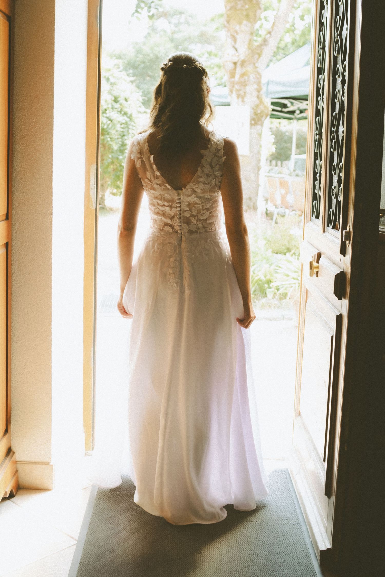 Bride in elegant lace-back white wedding gown standing in sunlit doorway, viewed from behind.