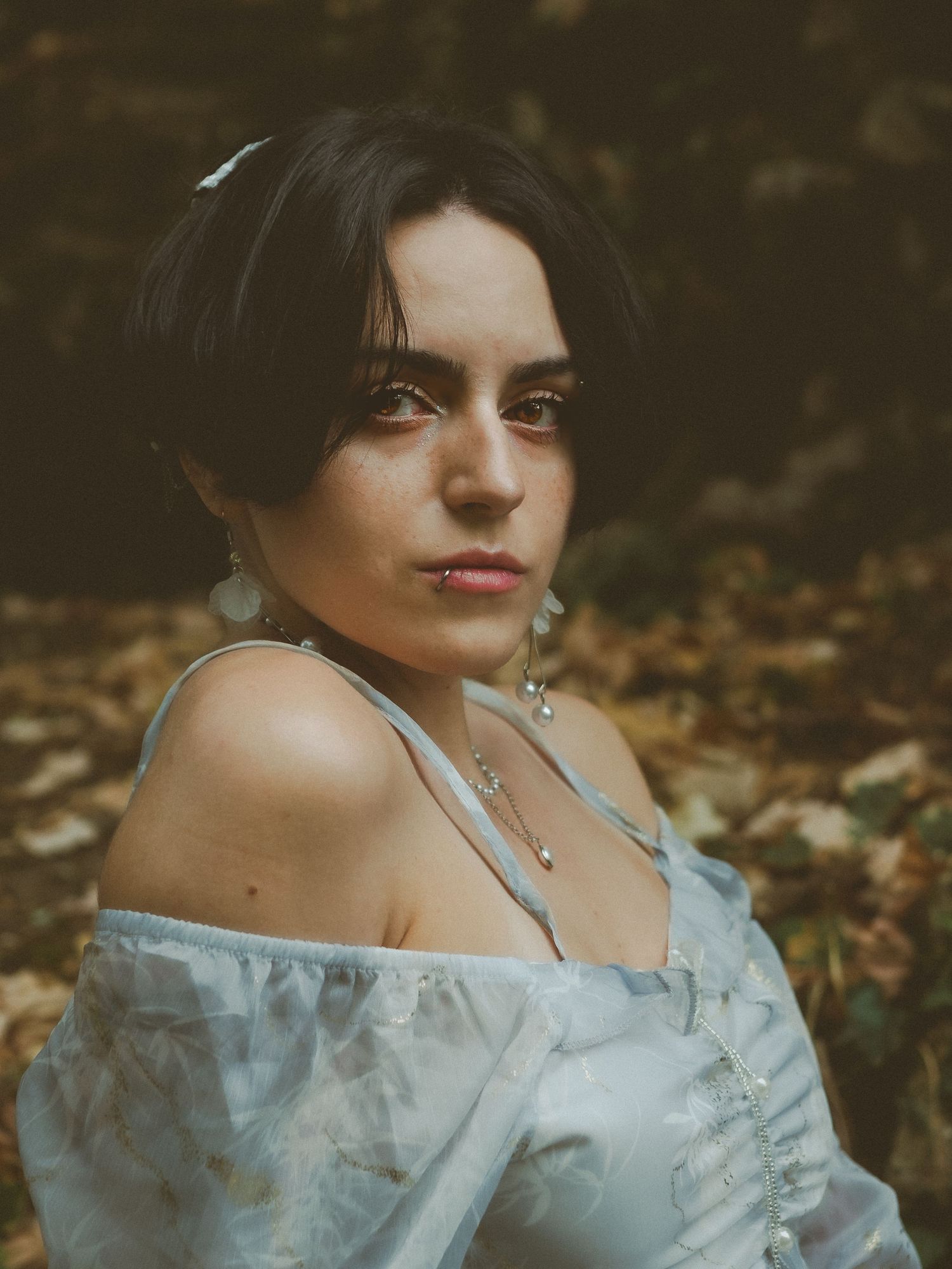 Woman in blue off-shoulder floral dress with dangling earrings poses outdoors among autumn leaves.