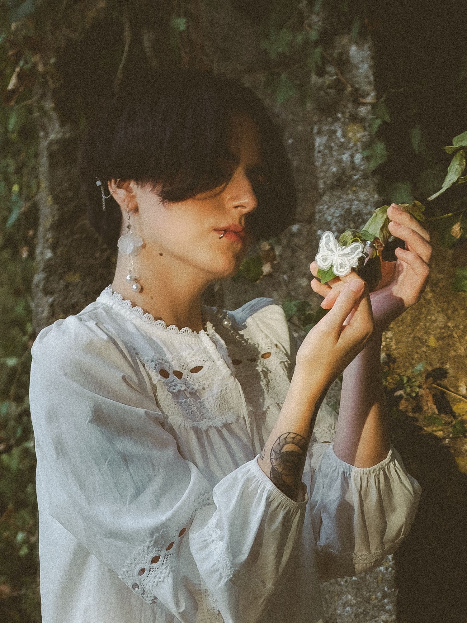 A woman in a white lace blouse holds delicate white flowers near mossy rocks, with a tattoo visible on her arm.