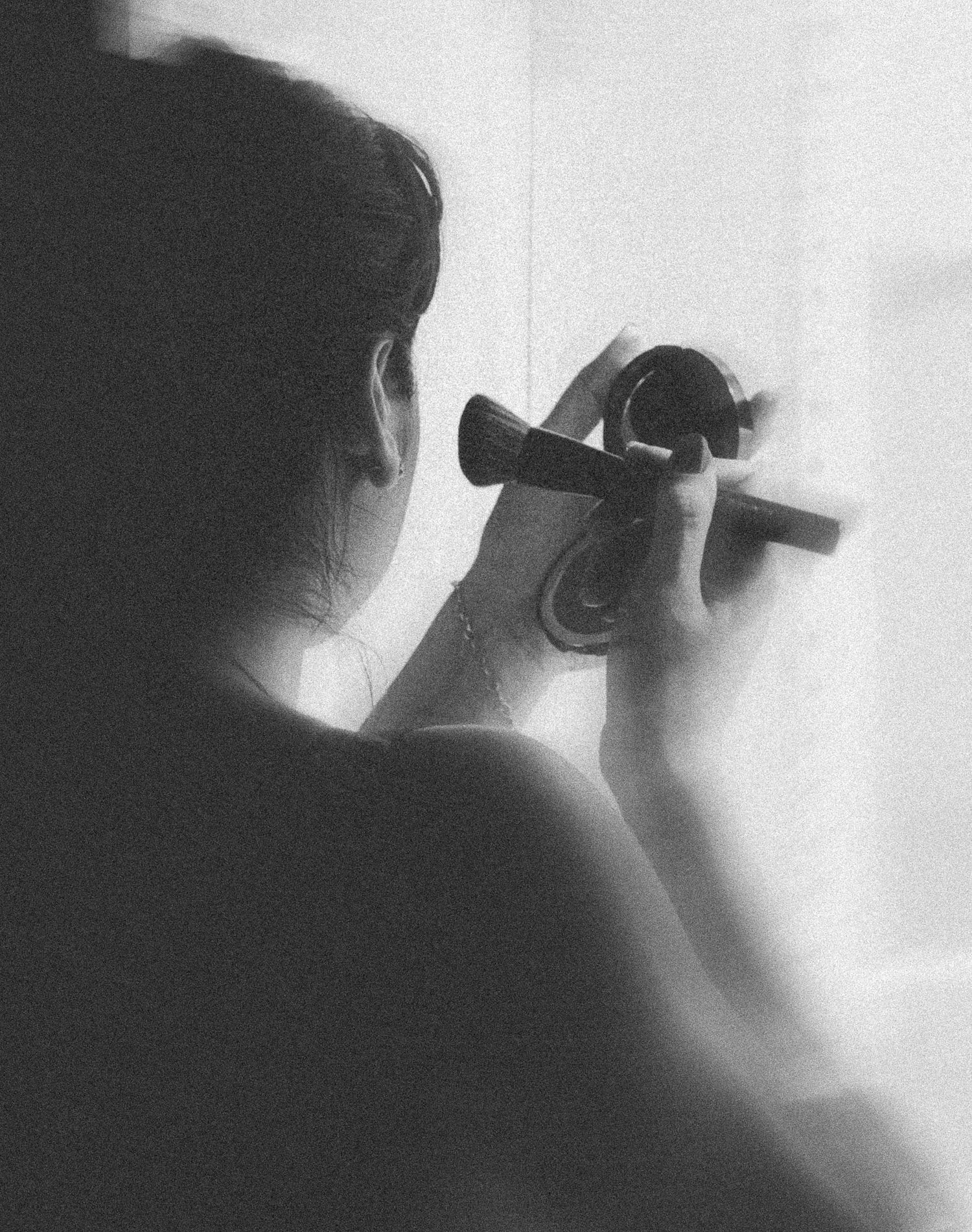 Vue de dos en noir et blanc d'une femme se maquillant devant un miroir près d'une fenêtre.