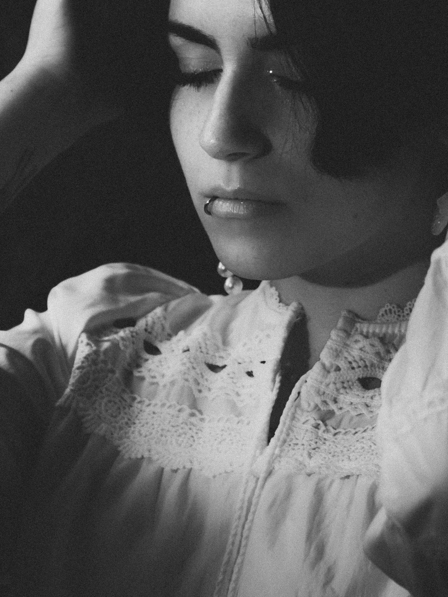 Black and white portrait of a young woman in lace blouse, eyes closed, hand raised near her head.