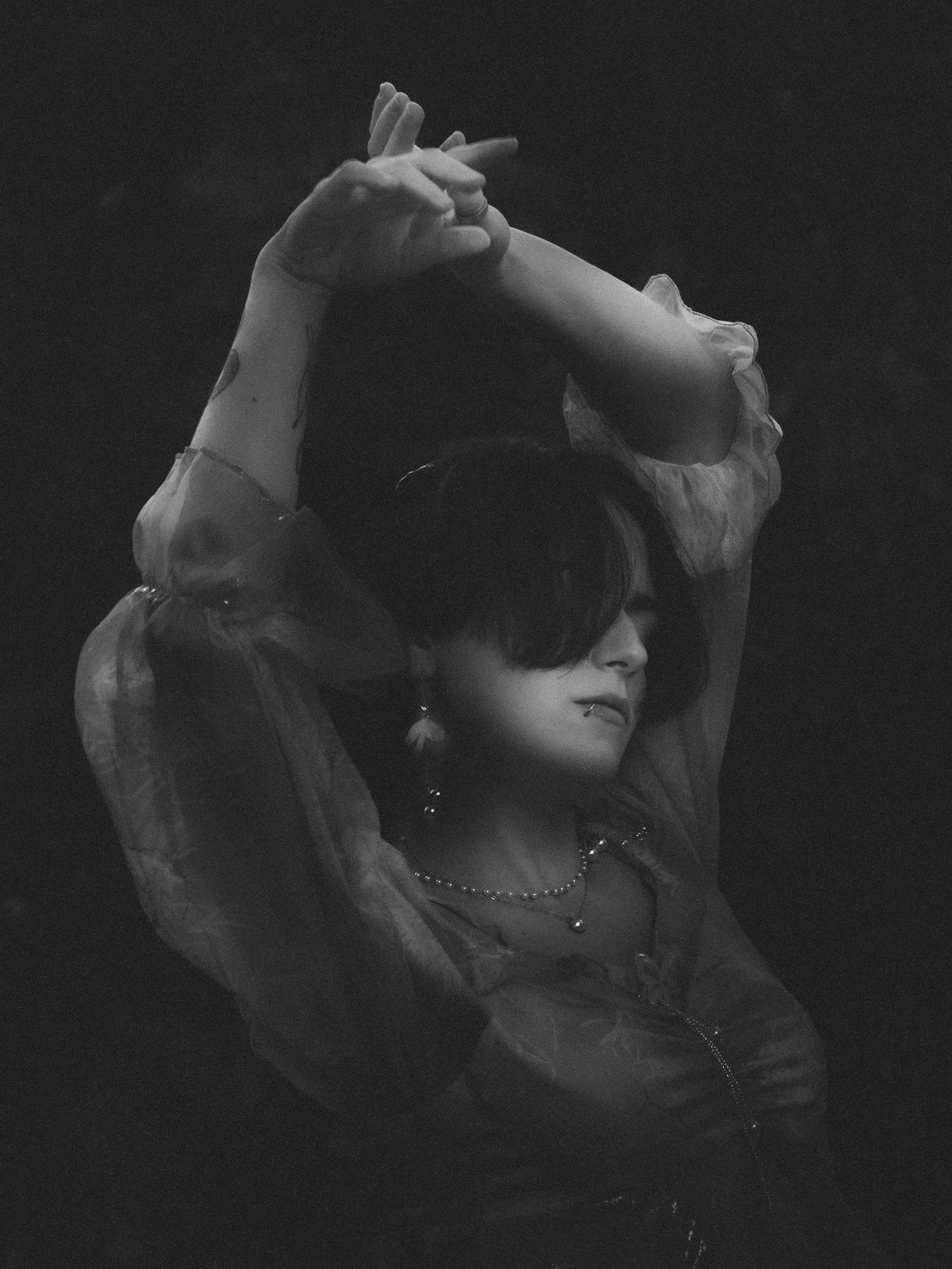 Black and white photo of a woman with arms raised gracefully overhead, wearing sheer blouse and delicate jewelry.
