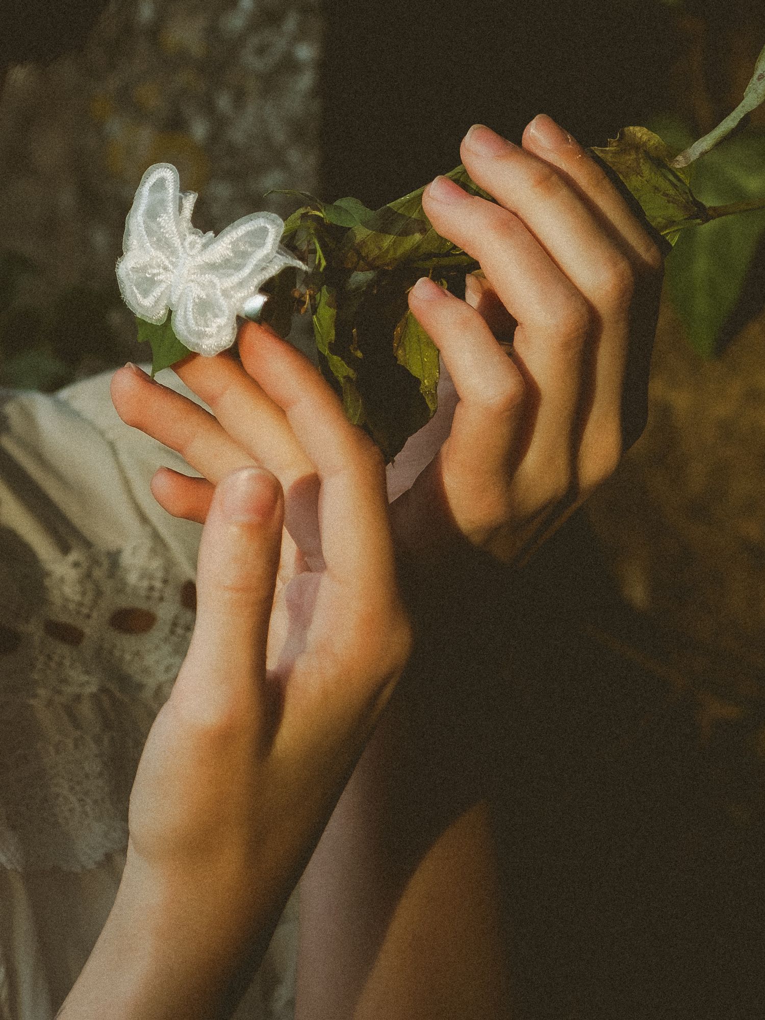 Delicate hands holding green leaves and a white butterfly ornament in soft natural light.