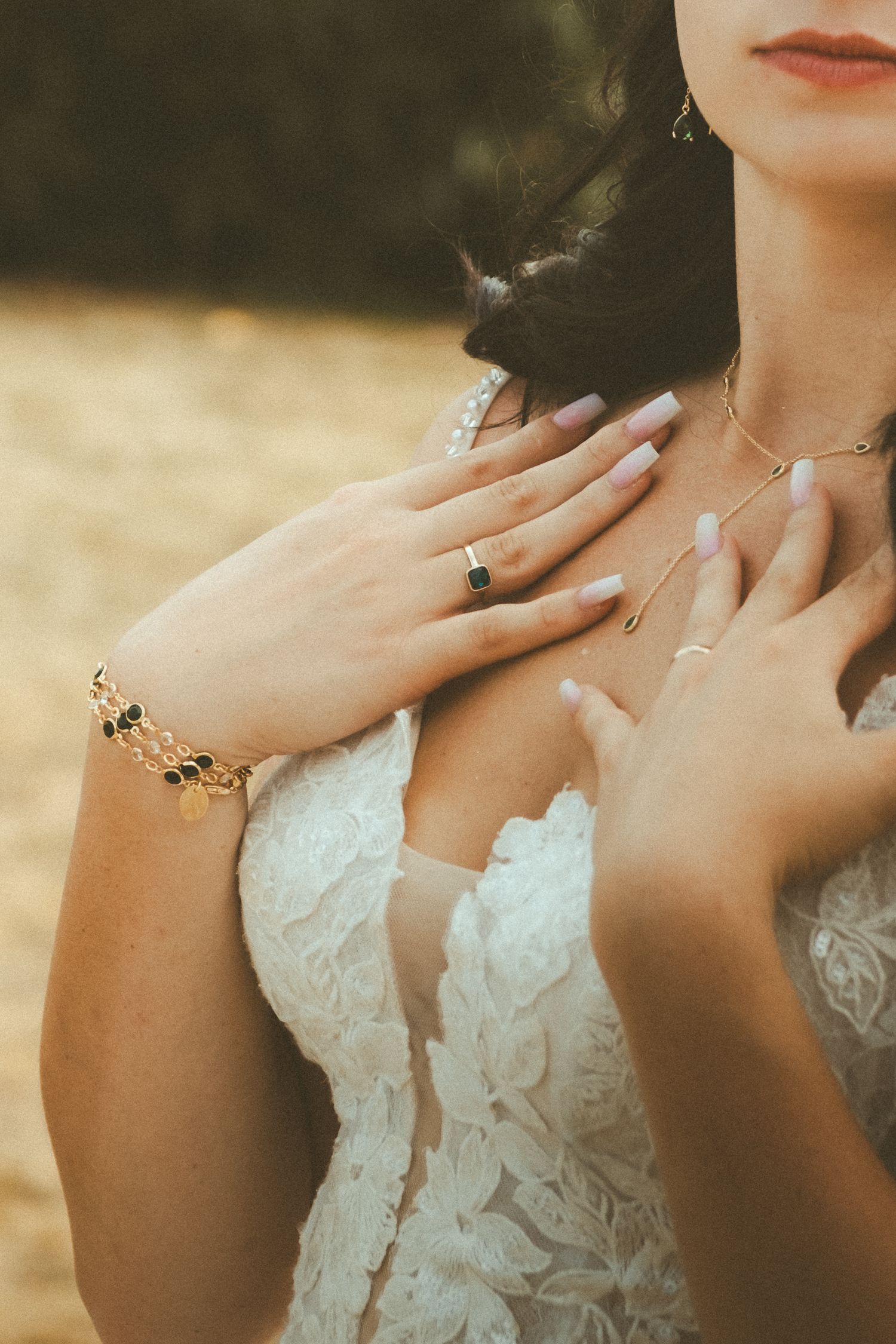 Bride in lace wedding dress with gold bracelet, emerald ring, and delicate necklace posing with hands on chest.