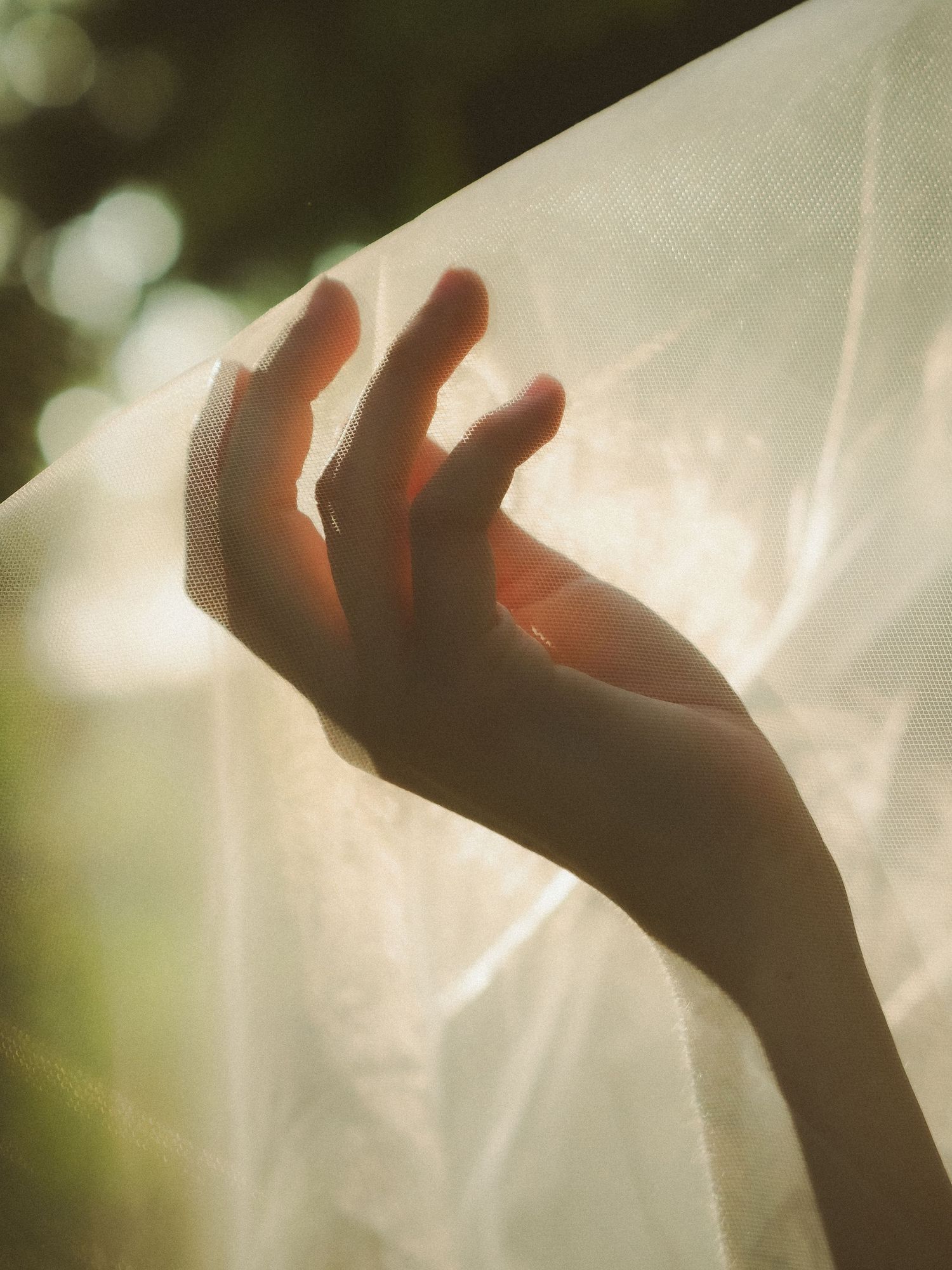 A hand gracefully reaches through sheer white fabric with soft bokeh green background.