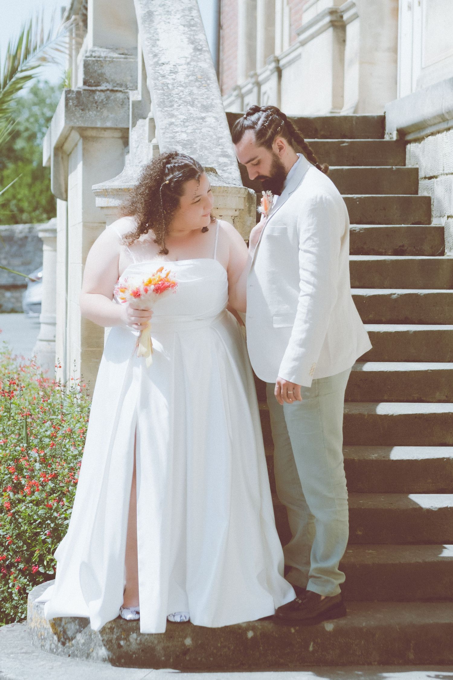 « Une mariée en robe blanche tenant son bouquet et son époux en veste blanche posant sur les marches en pierre d'un château.