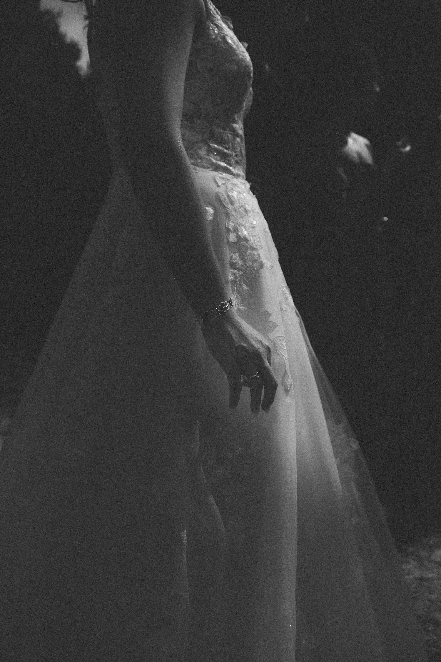 Black and white photo of a bride in a lace wedding gown, wearing a bracelet, standing elegantly in soft light.