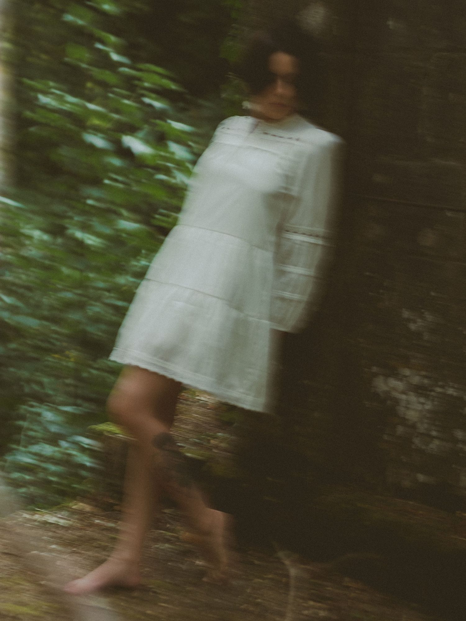 Woman in white tiered lace dress leaning against mossy stone wall surrounded by lush green foliage.