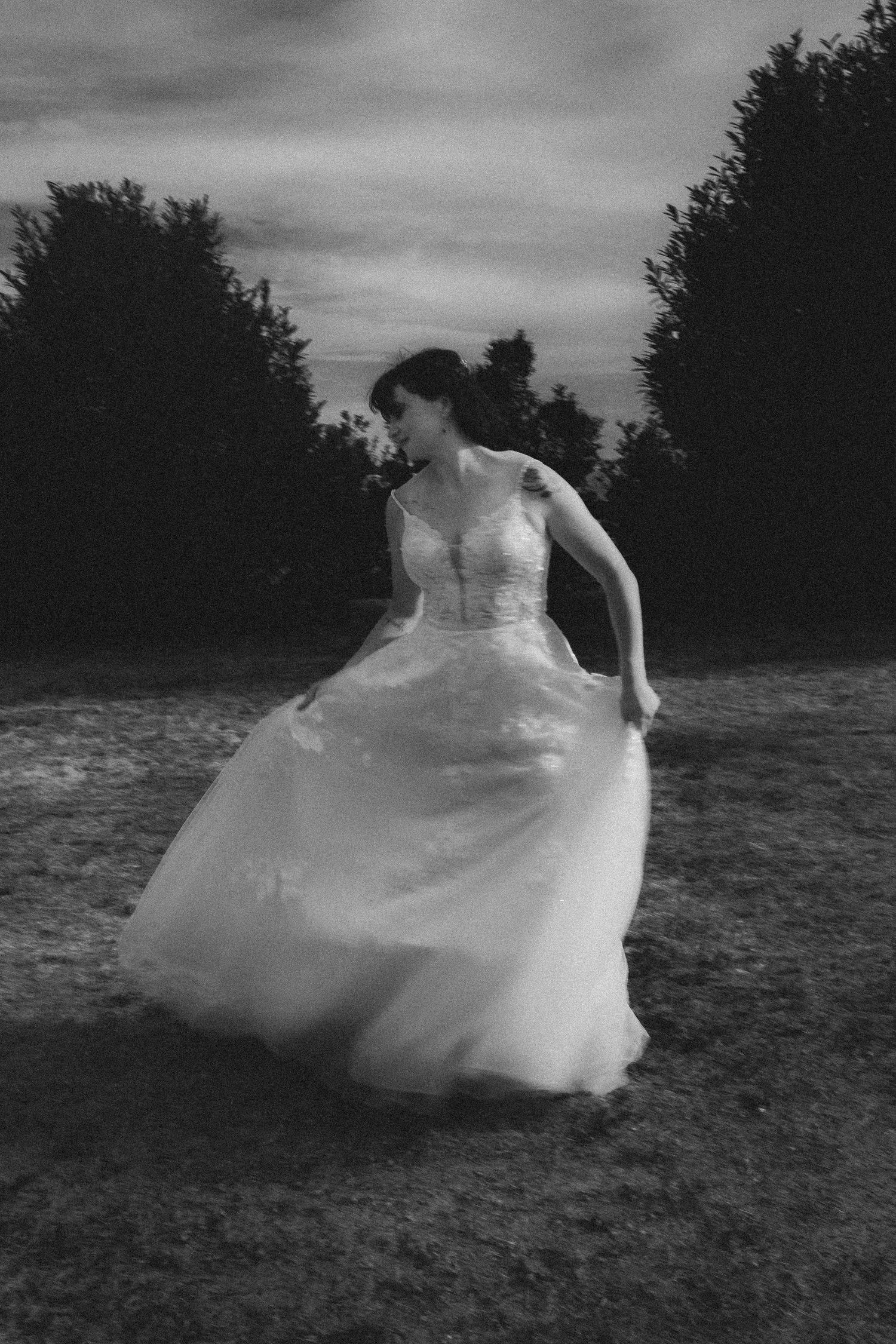 Black and white photo of a bride in a flowing wedding gown twirling outdoors near trees at dusk.
