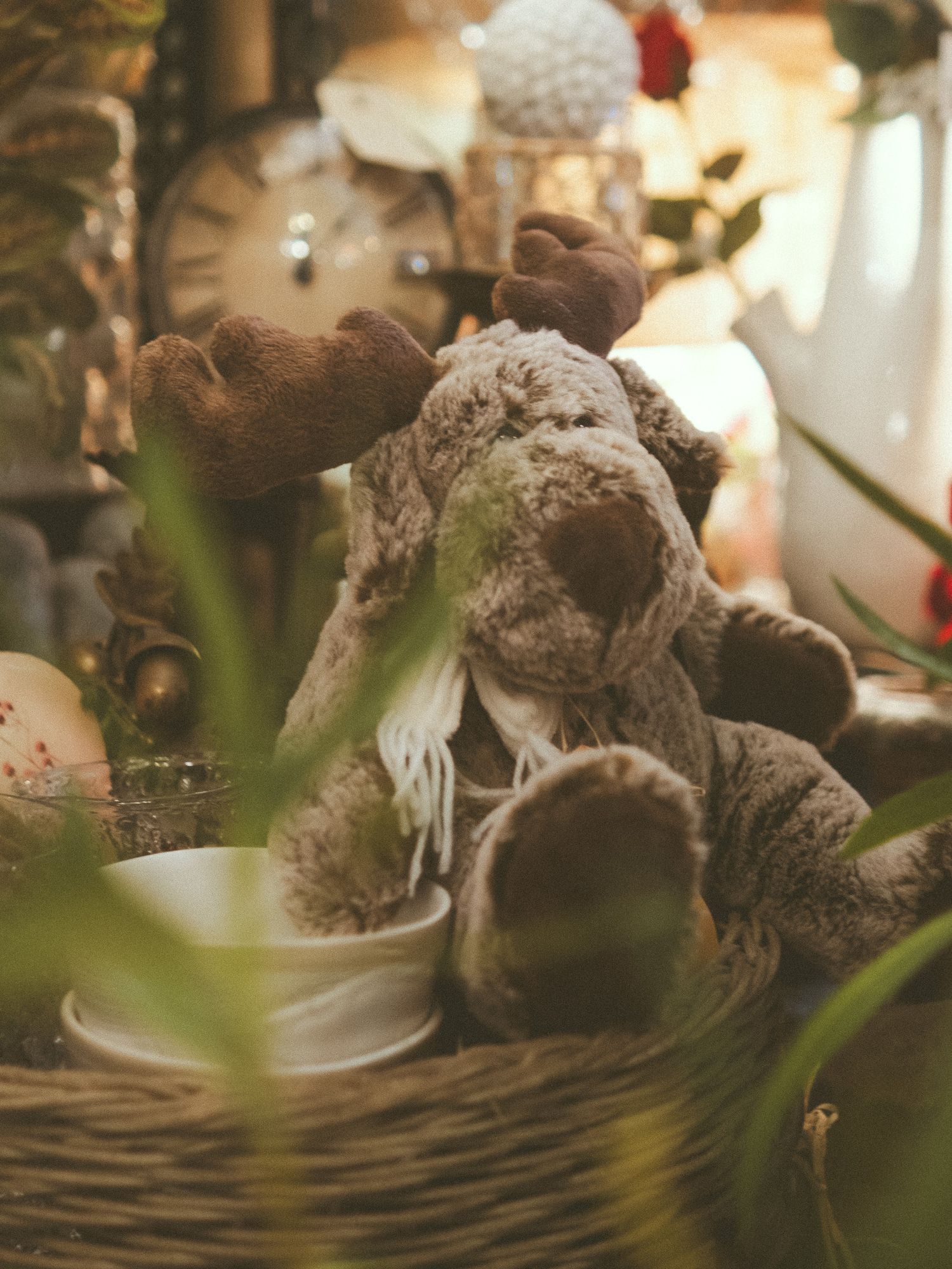 A cozy stuffed moose toy with a scarf sits in a wicker basket surrounded by festive Christmas decorations.