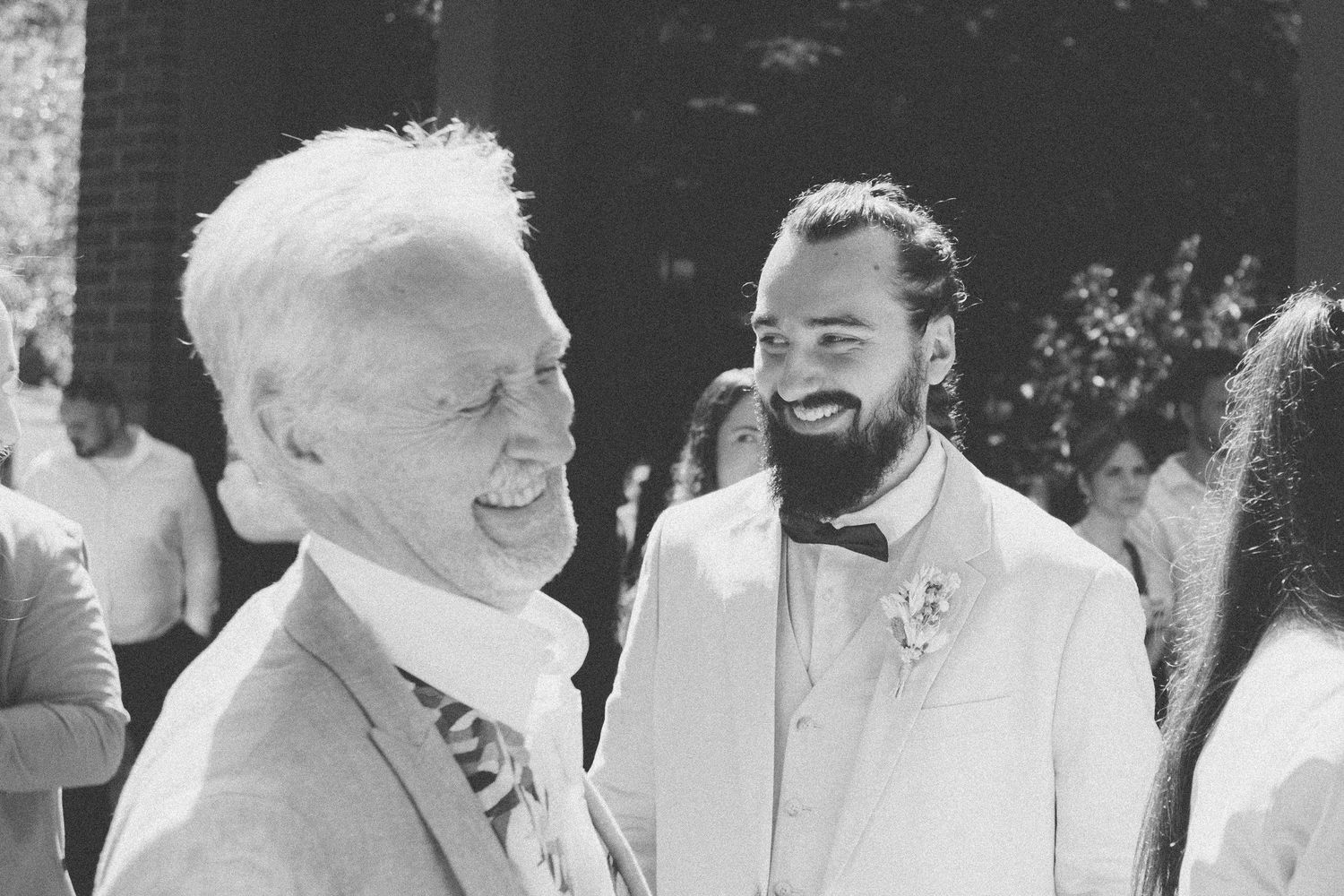 Un homme âgé et un marié barbu en costume blanc partagent un rire joyeux lors d'une cérémonie de mariage en extérieur.
