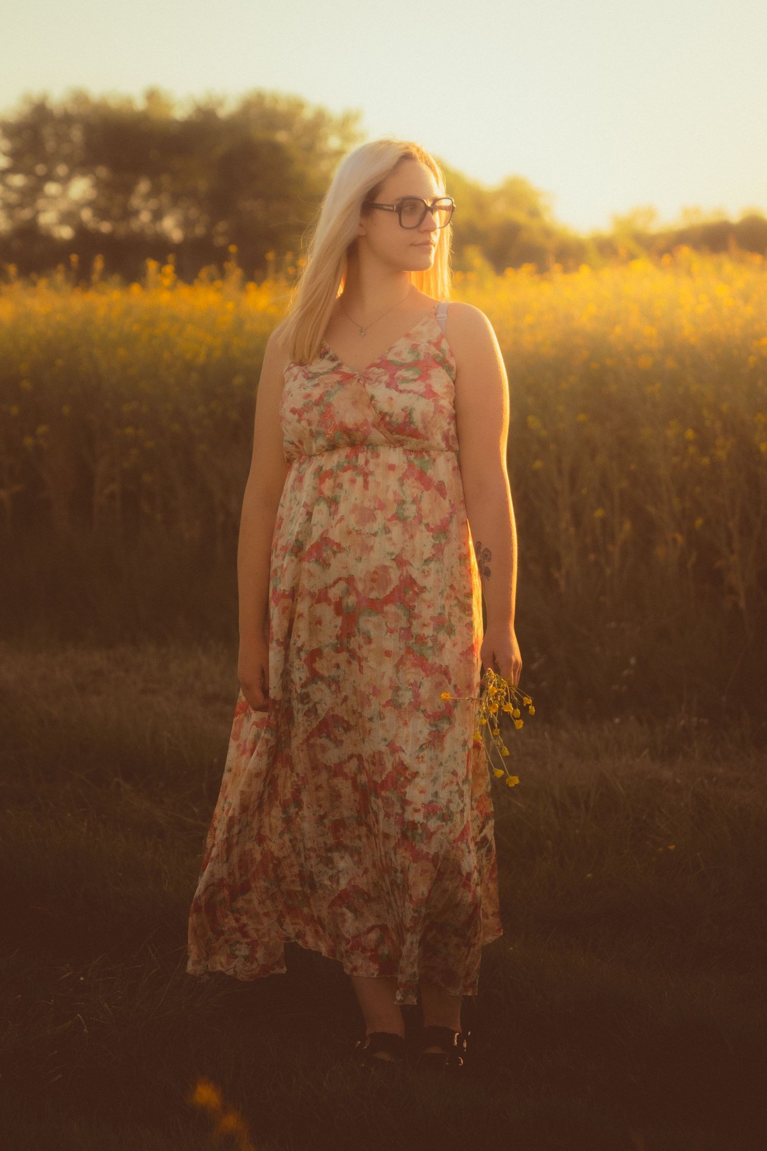 Blonde woman in floral maxi dress holding wildflowers in a golden sunset field.