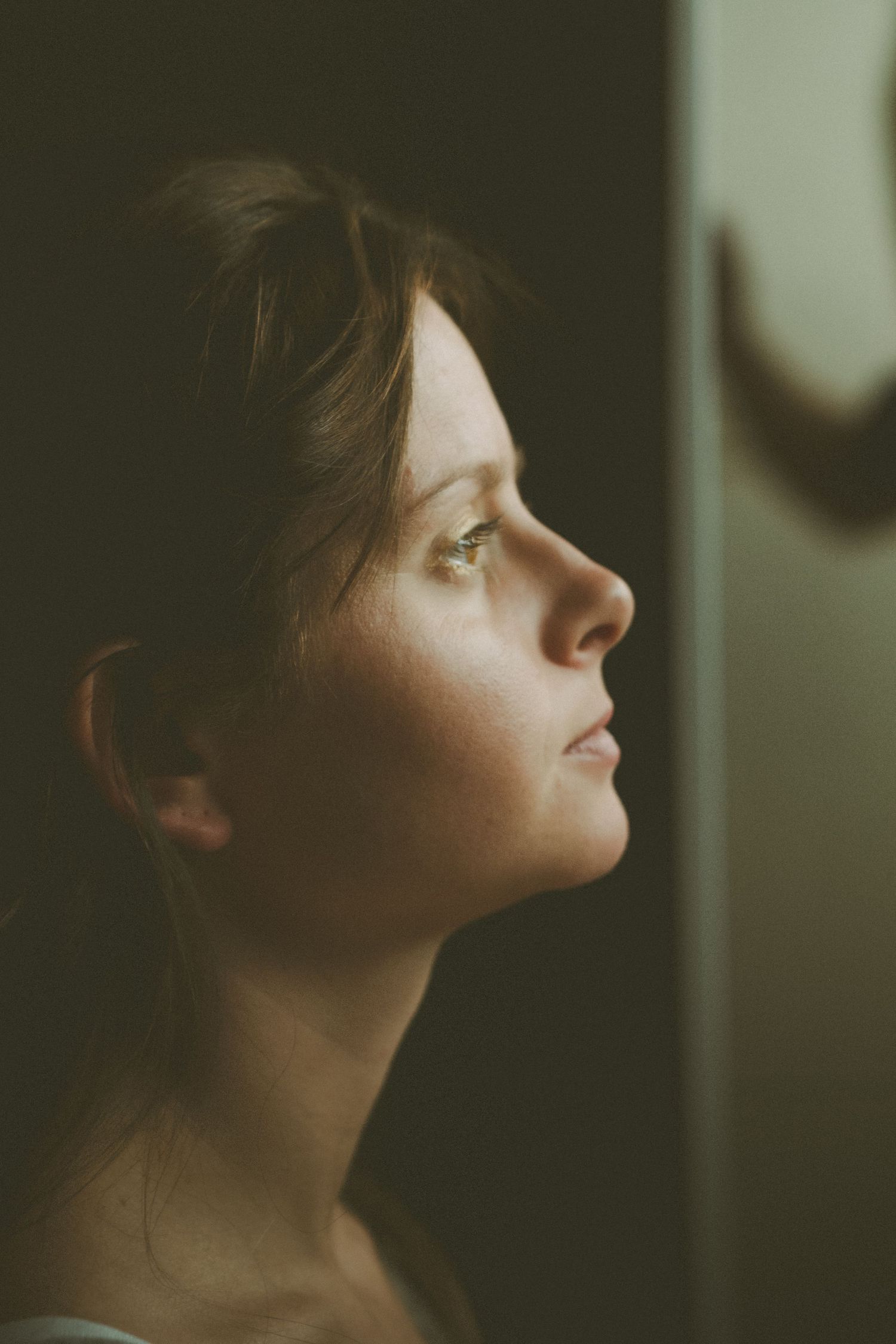 Side profile of a young woman with brown hair gazing upward in dramatic moody lighting.