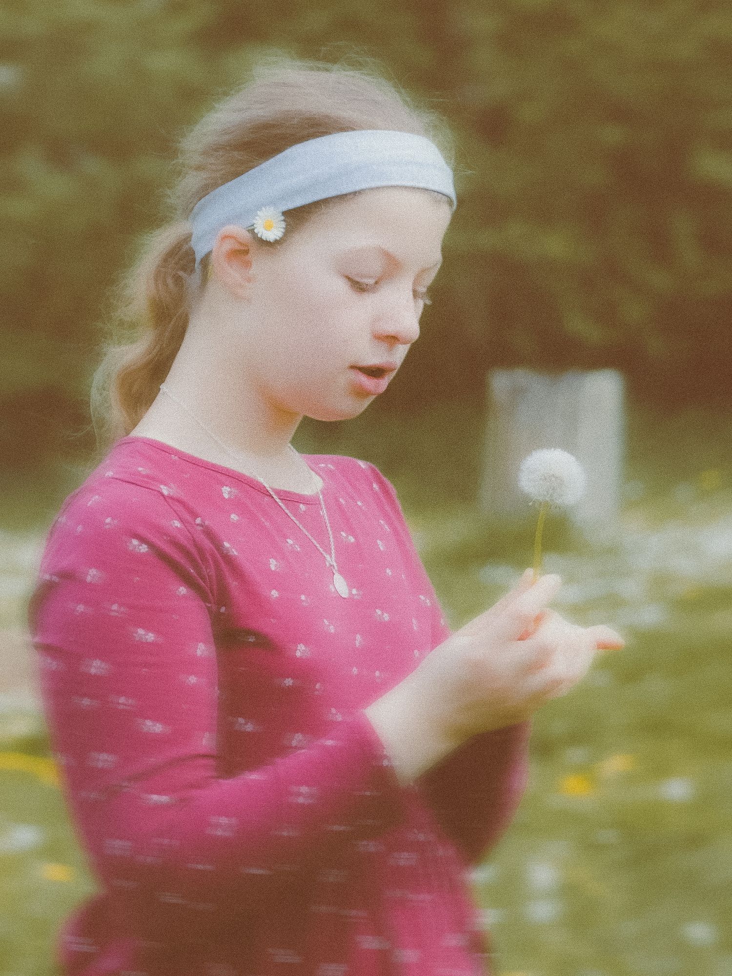 A young girl with a blue headband blows a dandelion wishflower in a soft-focus outdoor setting.