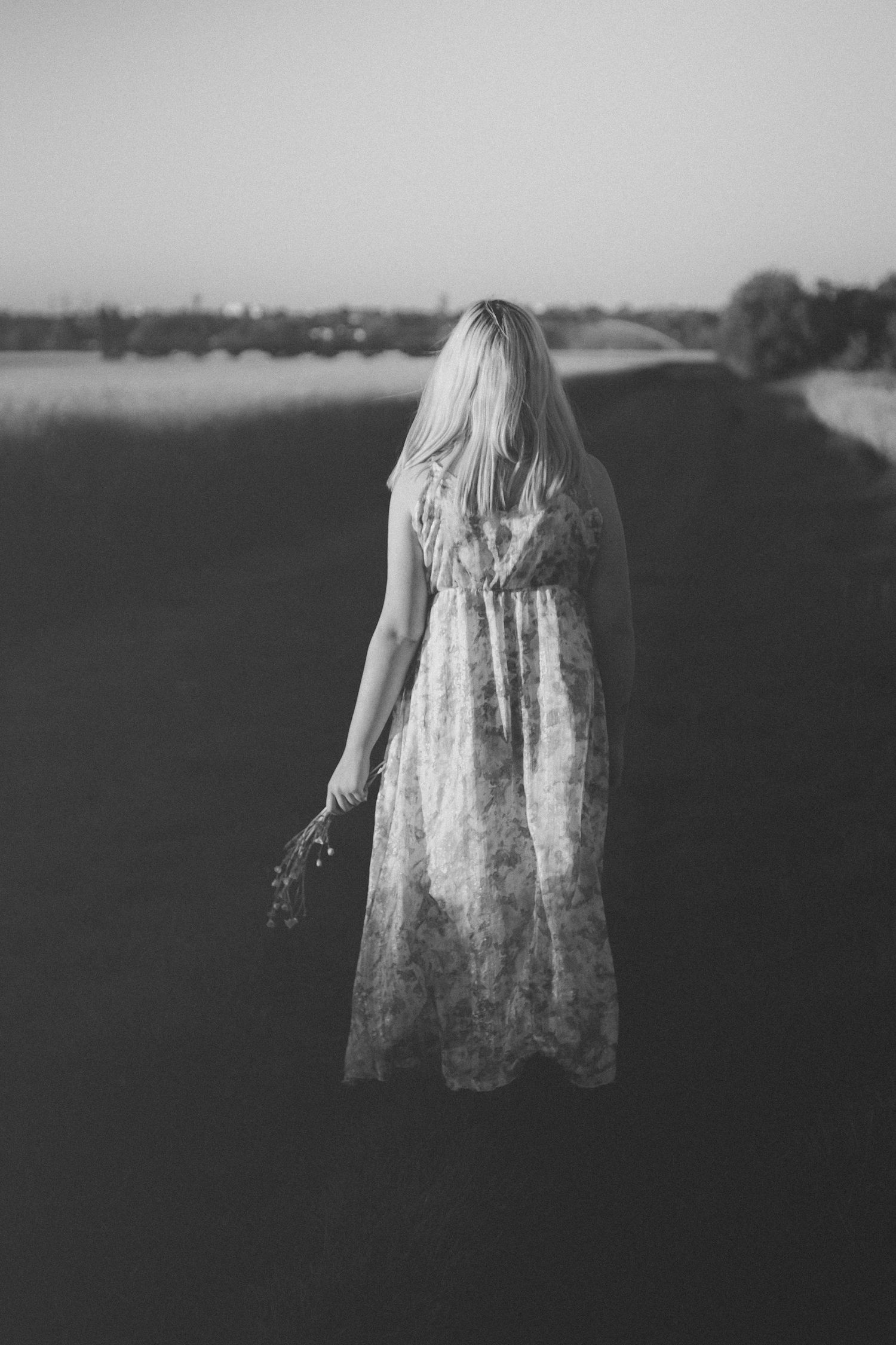 Black and white photo of a blonde woman in a floral dress holding wildflowers, standing alone in an open field.