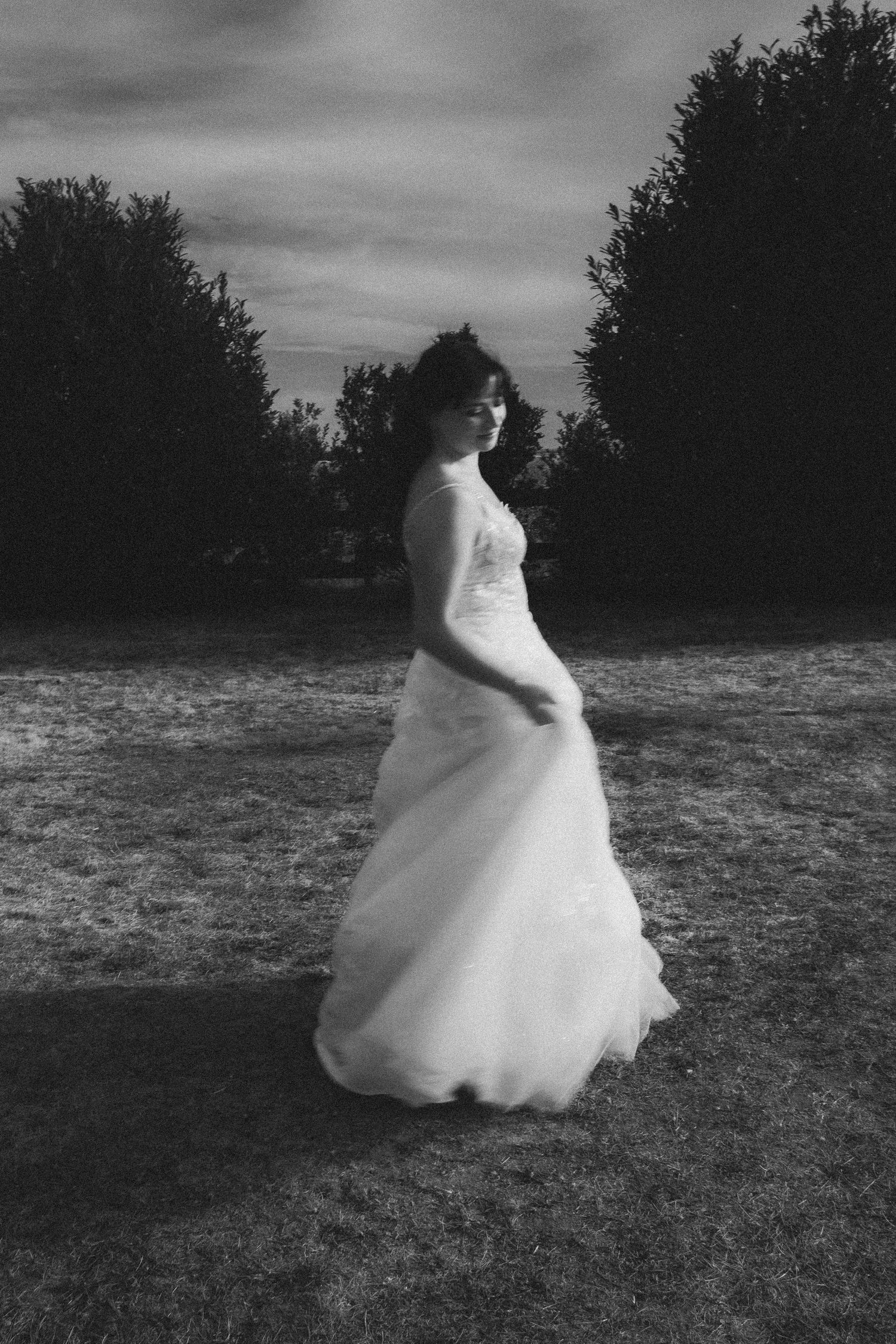 Black and white photo of a bride in a flowing white gown standing outdoors on grass with trees in the background.