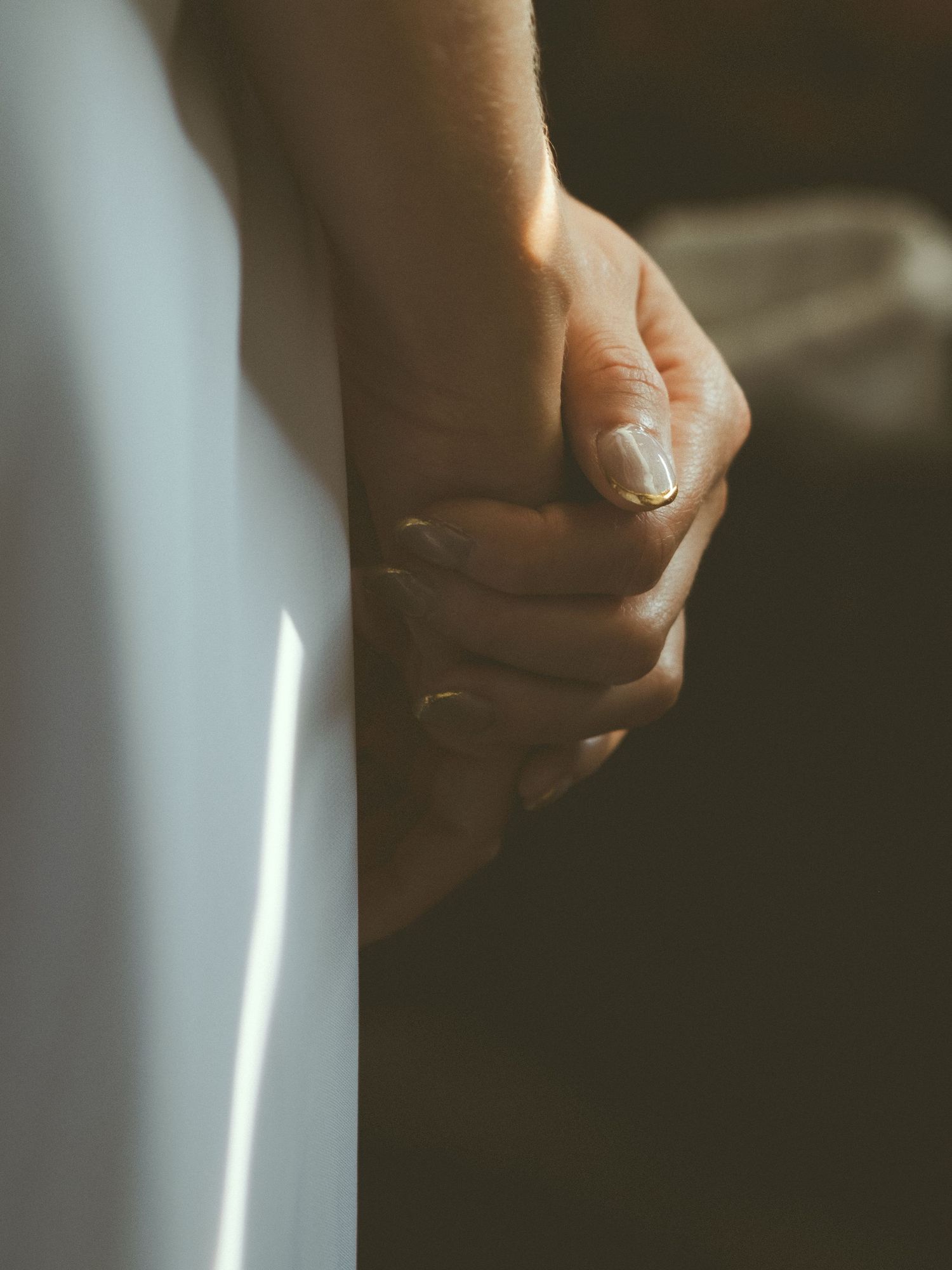Two hands holding together with a gold ring, bathed in soft natural light against a dark background.