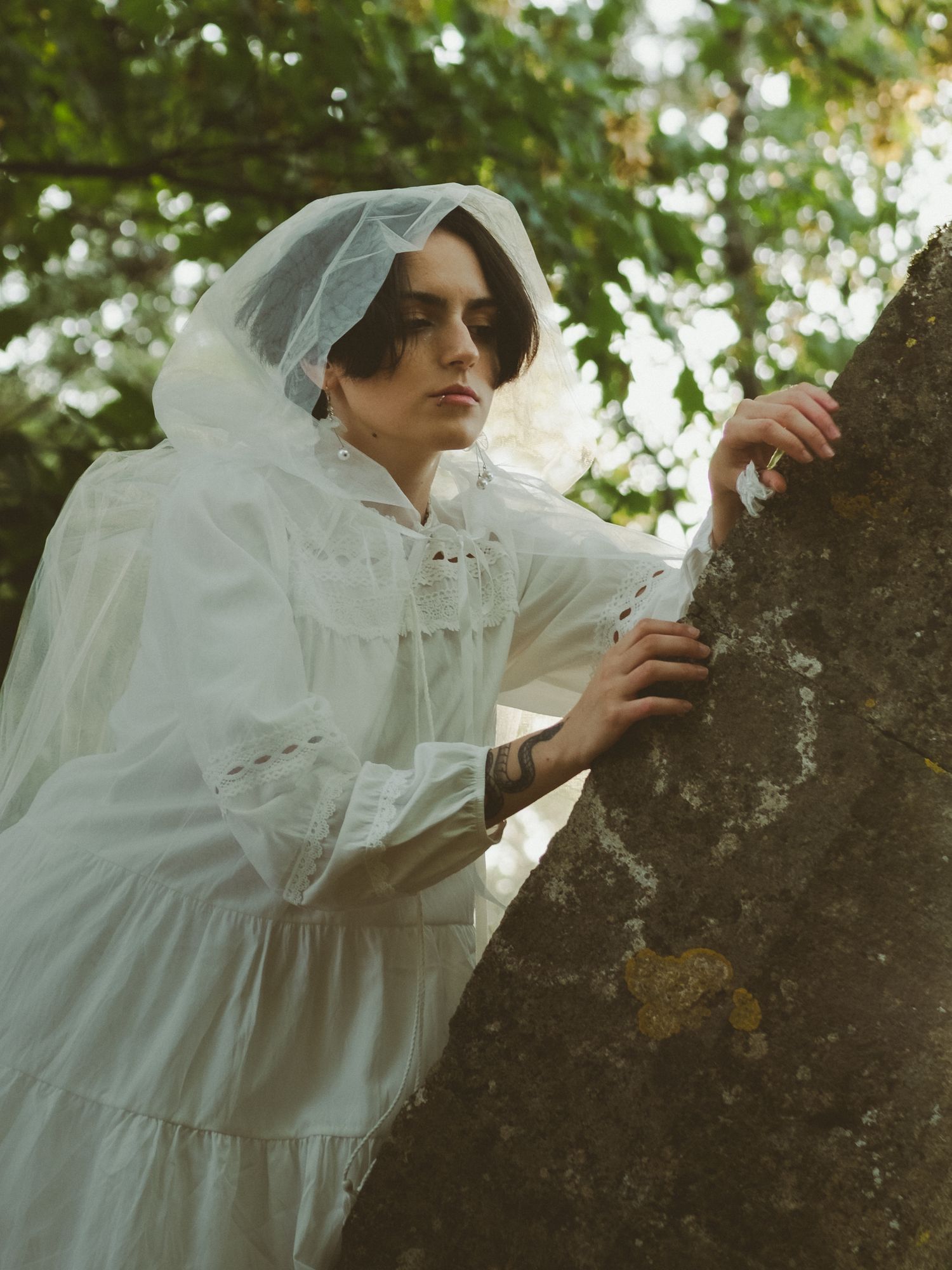A woman in a white Victorian-style dress and veil leans against a lichen-covered stone outdoors.