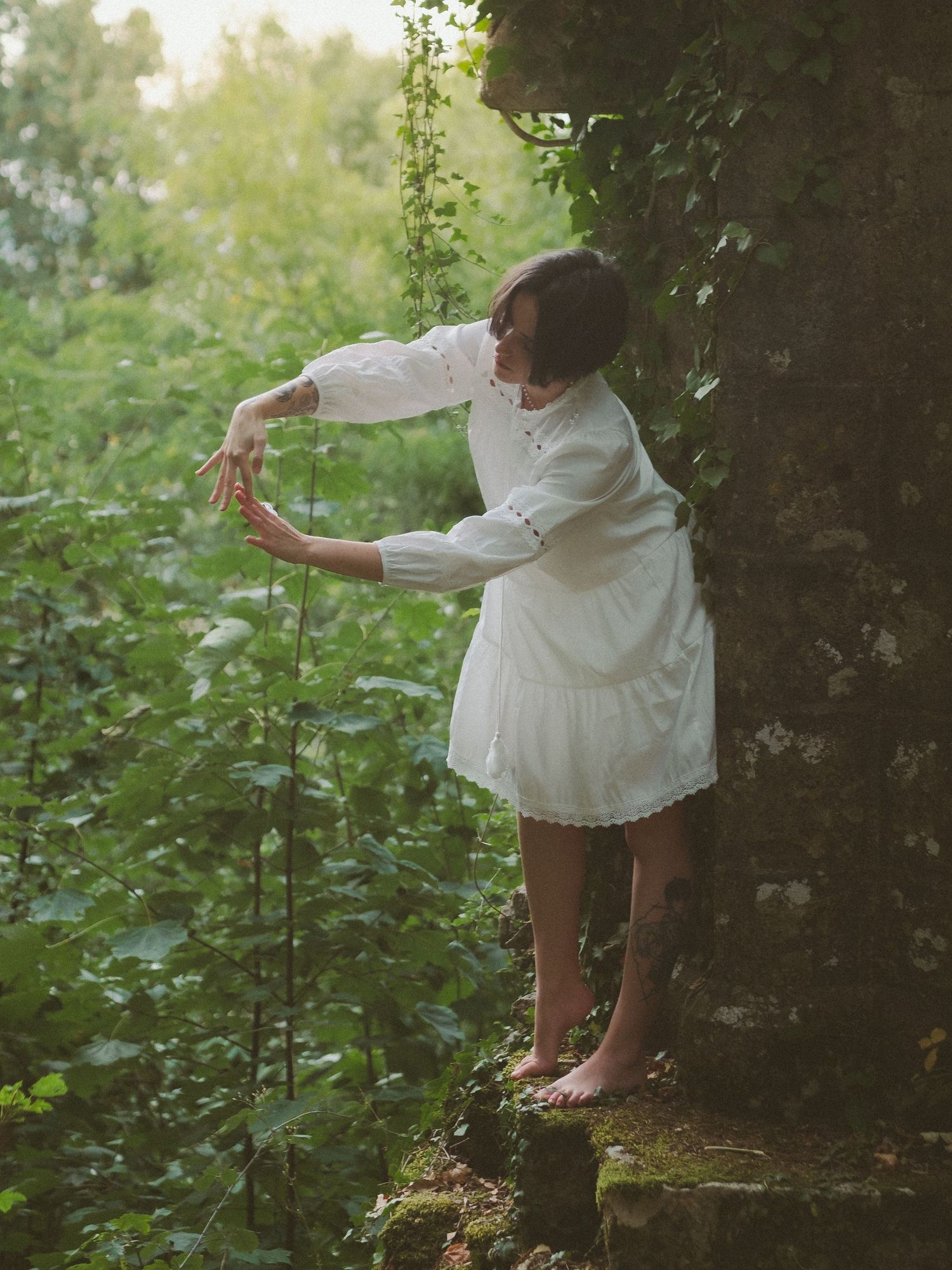 Barefoot woman in white dress reaching for ivy vines beside mossy stone wall in lush green garden.