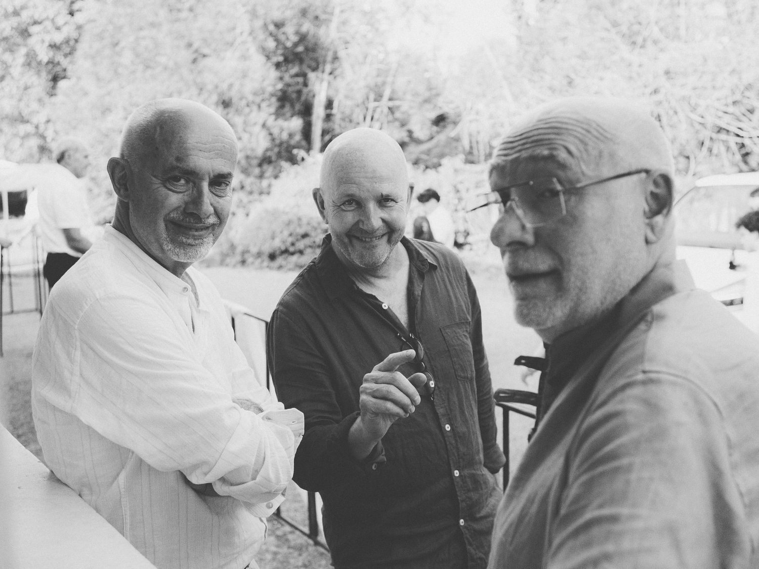 Photo en noir et blanc de trois hommes âgés souriant et discutant ensemble lors d'un mariage en extérieur.