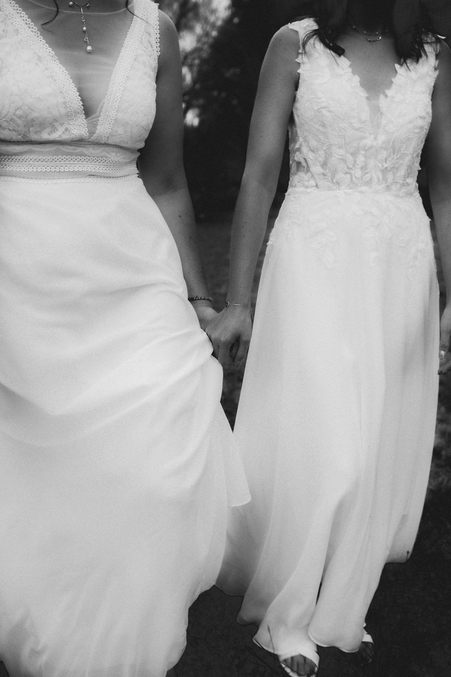 Two brides in white wedding gowns holding hands in a romantic black and white outdoor photo.