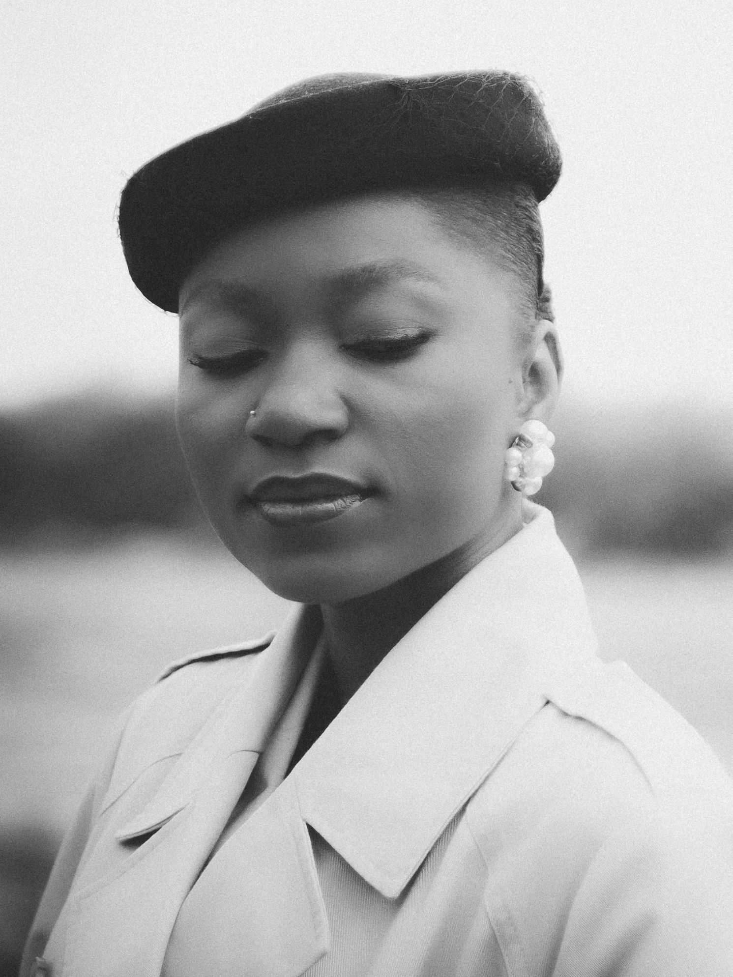 Black and white portrait of a woman wearing a beret and trench coat, with a nose ring and earrings.