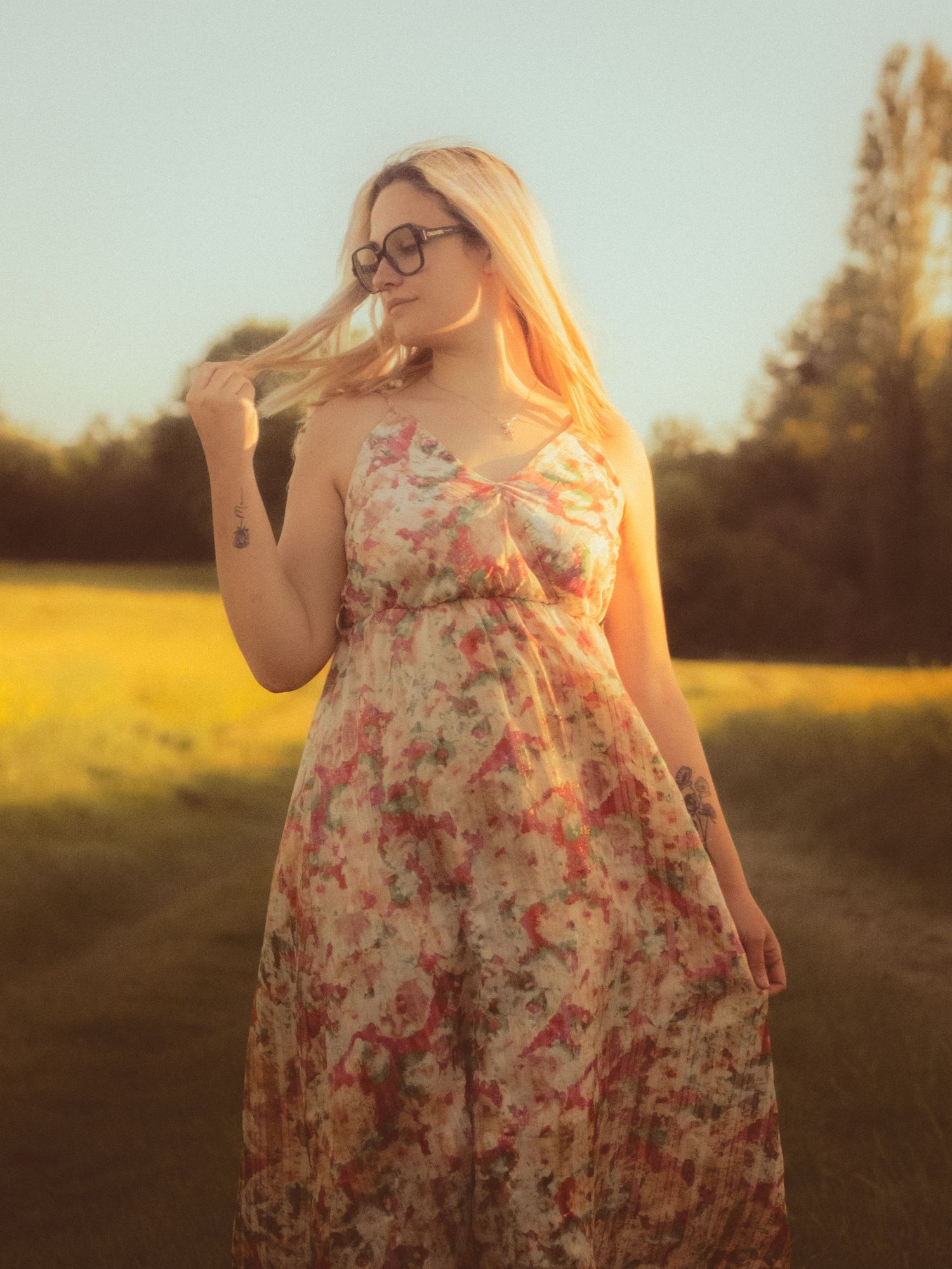 Blonde woman in floral sundress and glasses standing in golden hour sunlight in a scenic field.