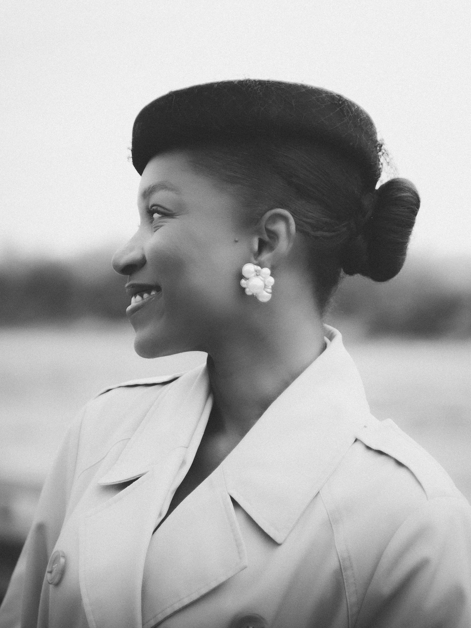 Black and white portrait of a smiling woman wearing a beret, floral earrings, and a trench coat.