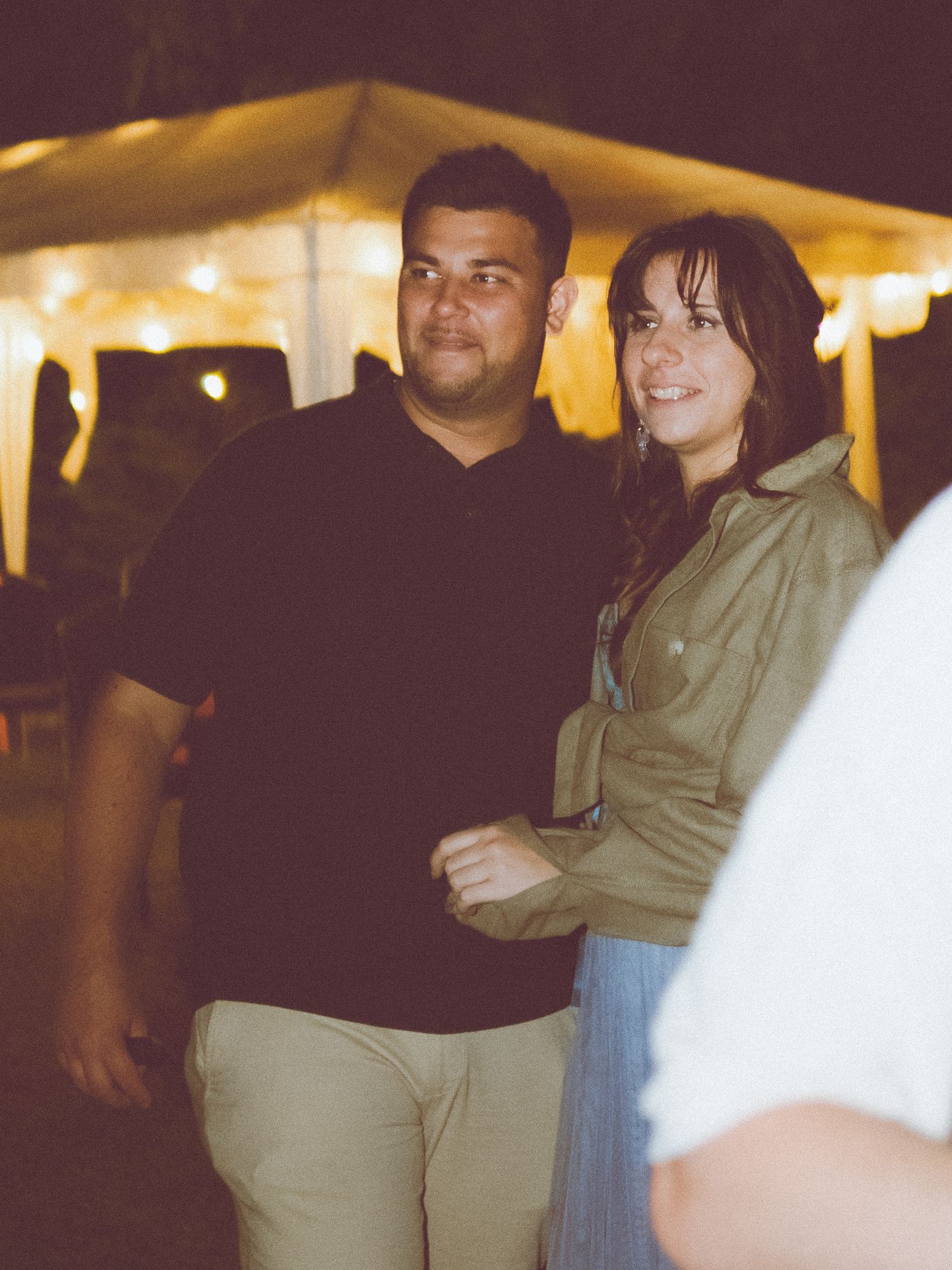 A couple stands together at an outdoor evening event with string lights illuminating a tent in the background.