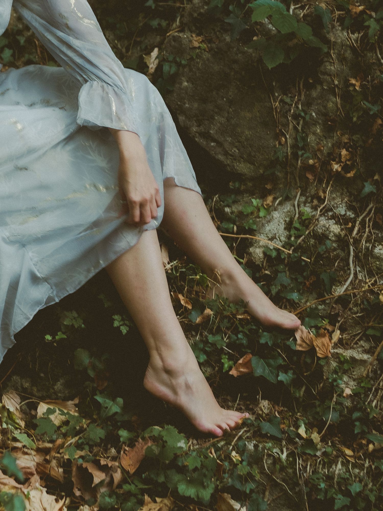 Barefoot woman in light blue dress sitting on forest floor surrounded by ivy and autumn leaves.