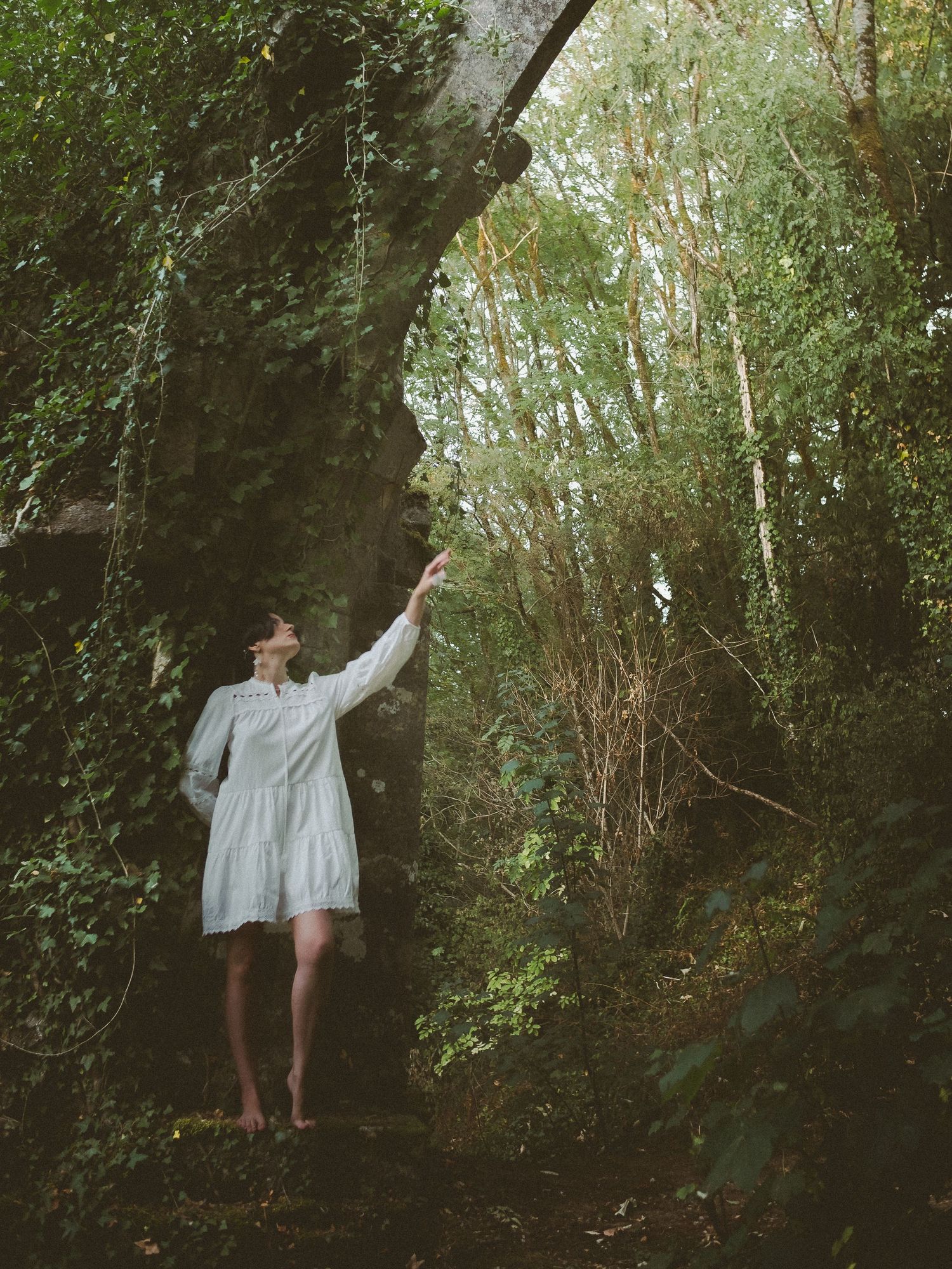 Woman in white dress touching mossy rock arch in lush green forest, barefoot among ivy and trees.