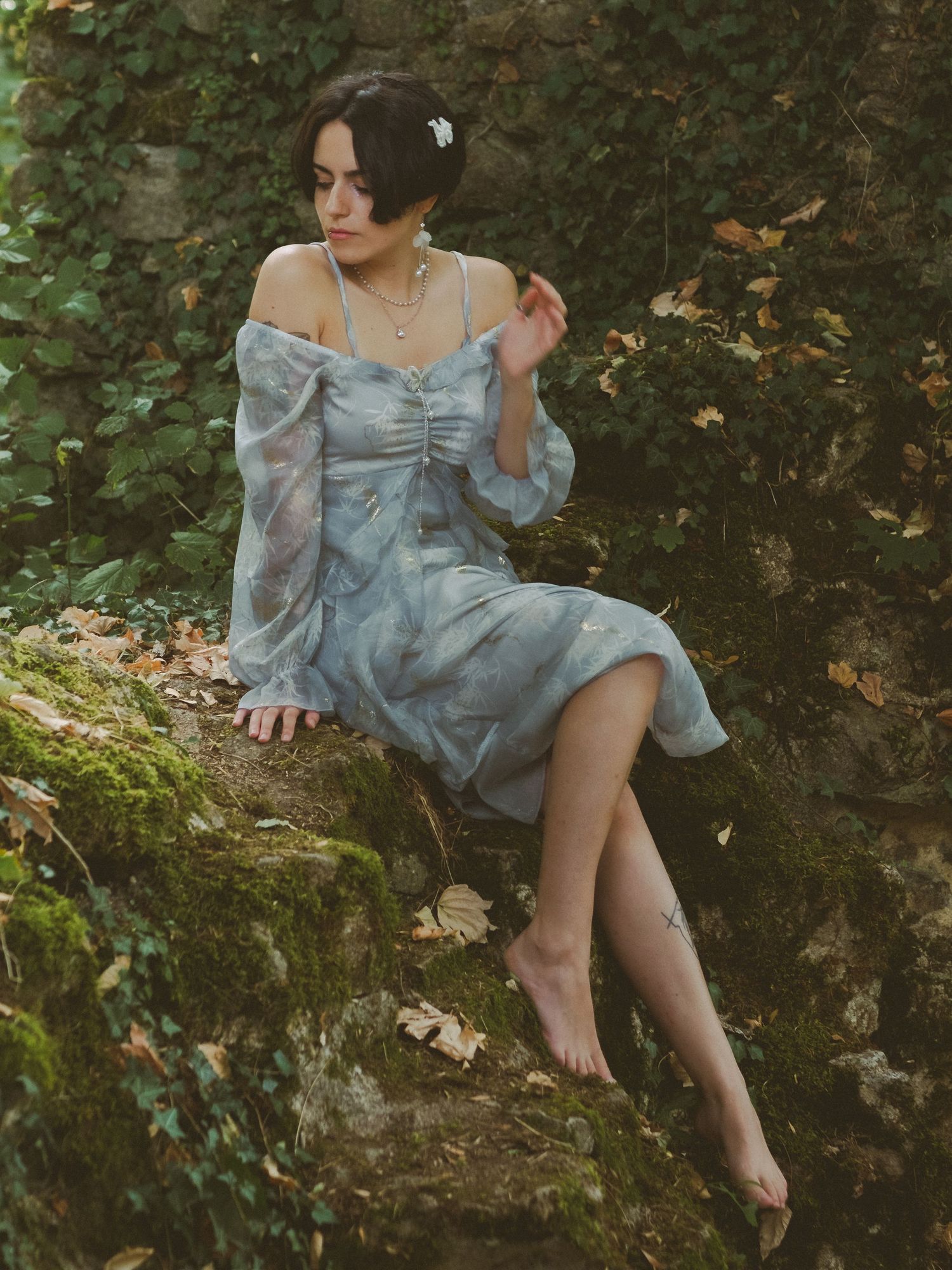Barefoot woman in floral blue off-shoulder dress sits gracefully on mossy stone wall.