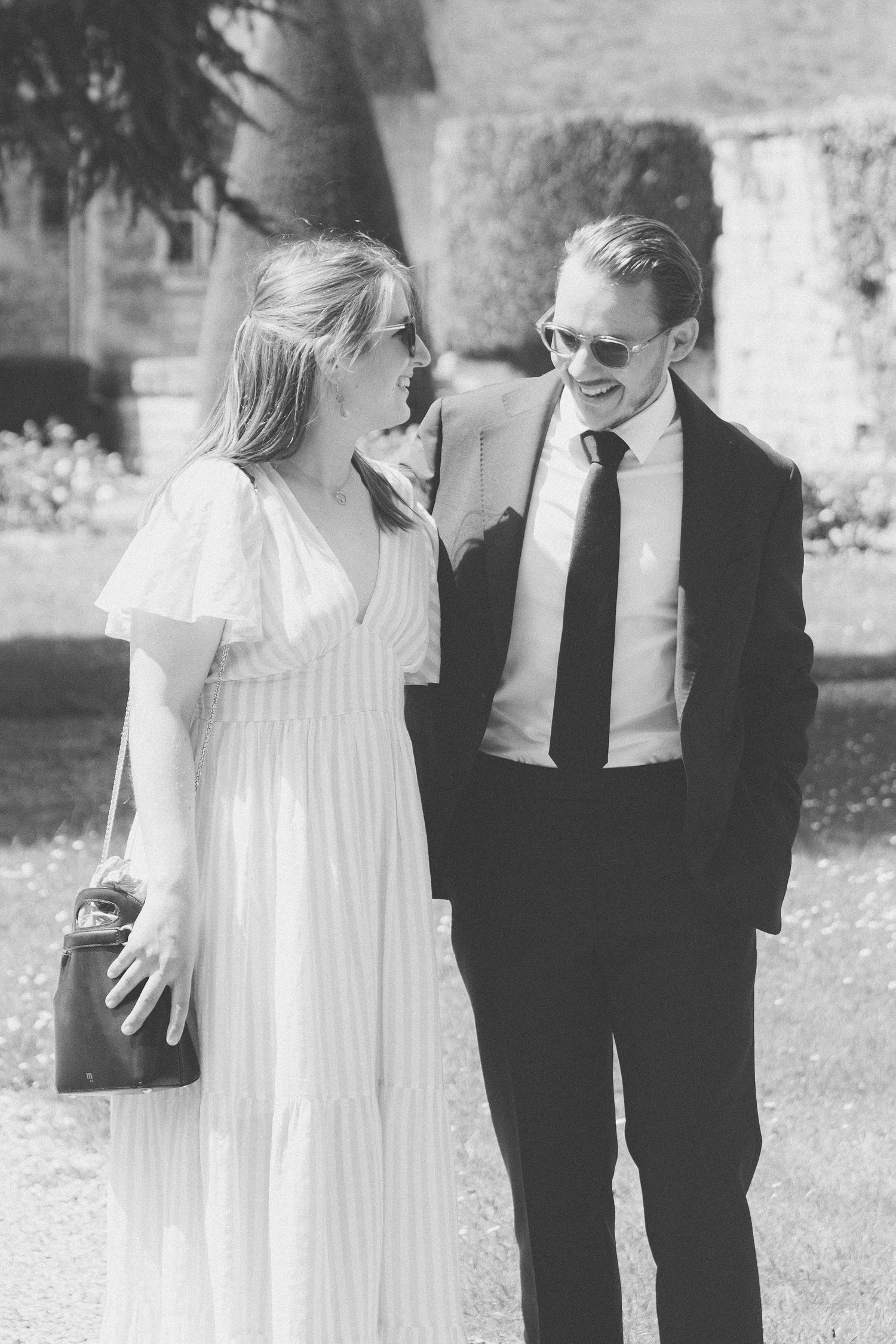Black and white photo of an elegant couple in formal attire walking together outdoors at a garden event.