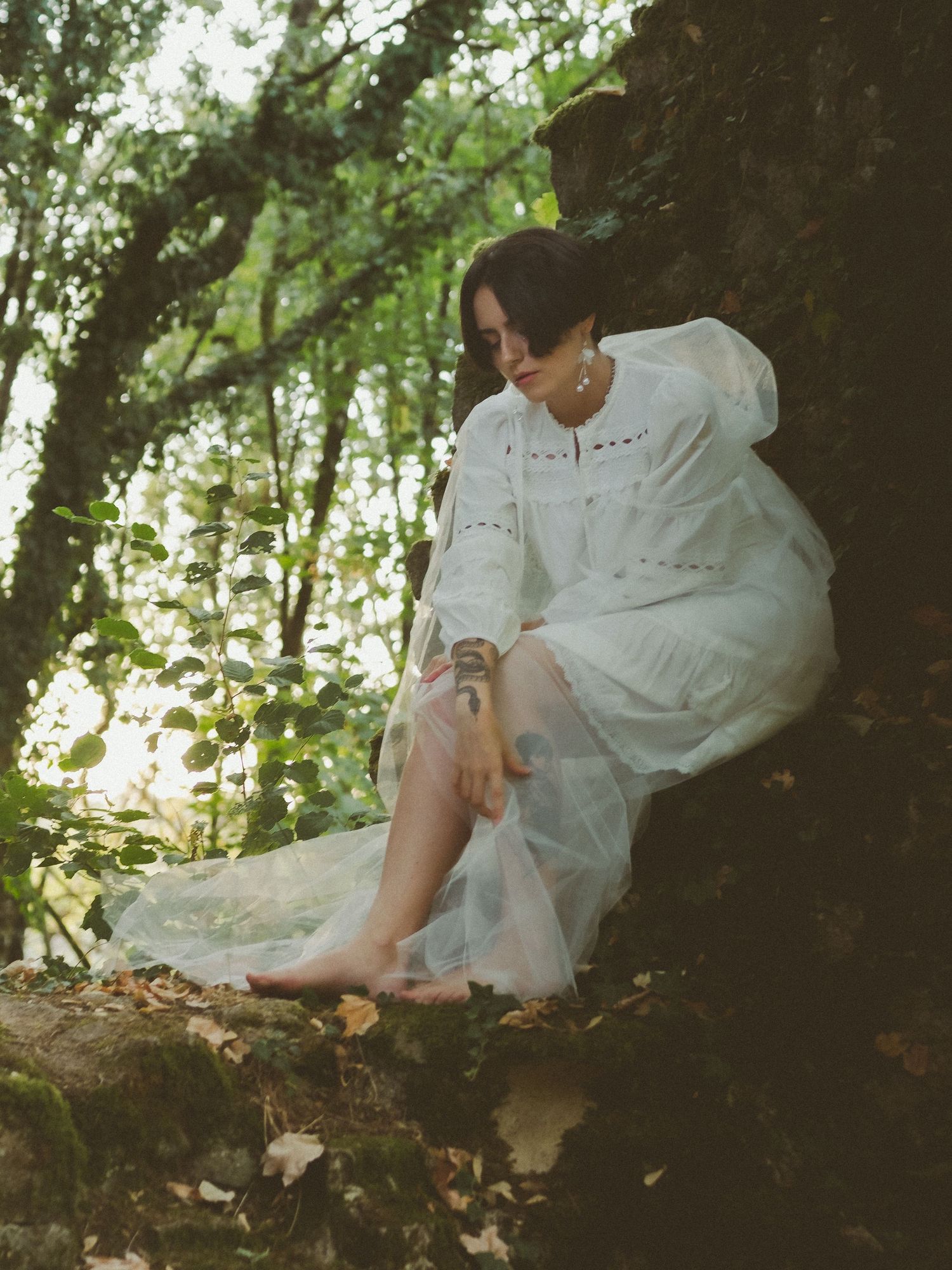 Woman in white dress sitting barefoot against mossy rocks in a lush green forest setting.