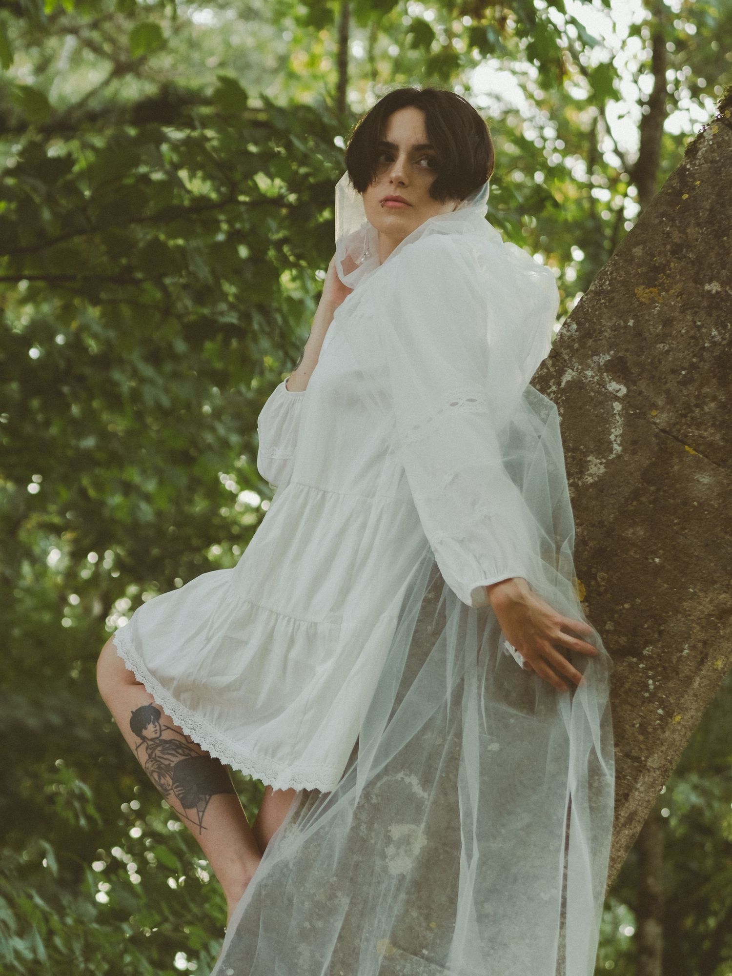 Woman in white sheer tiered dress leans against tree surrounded by lush green foliage.