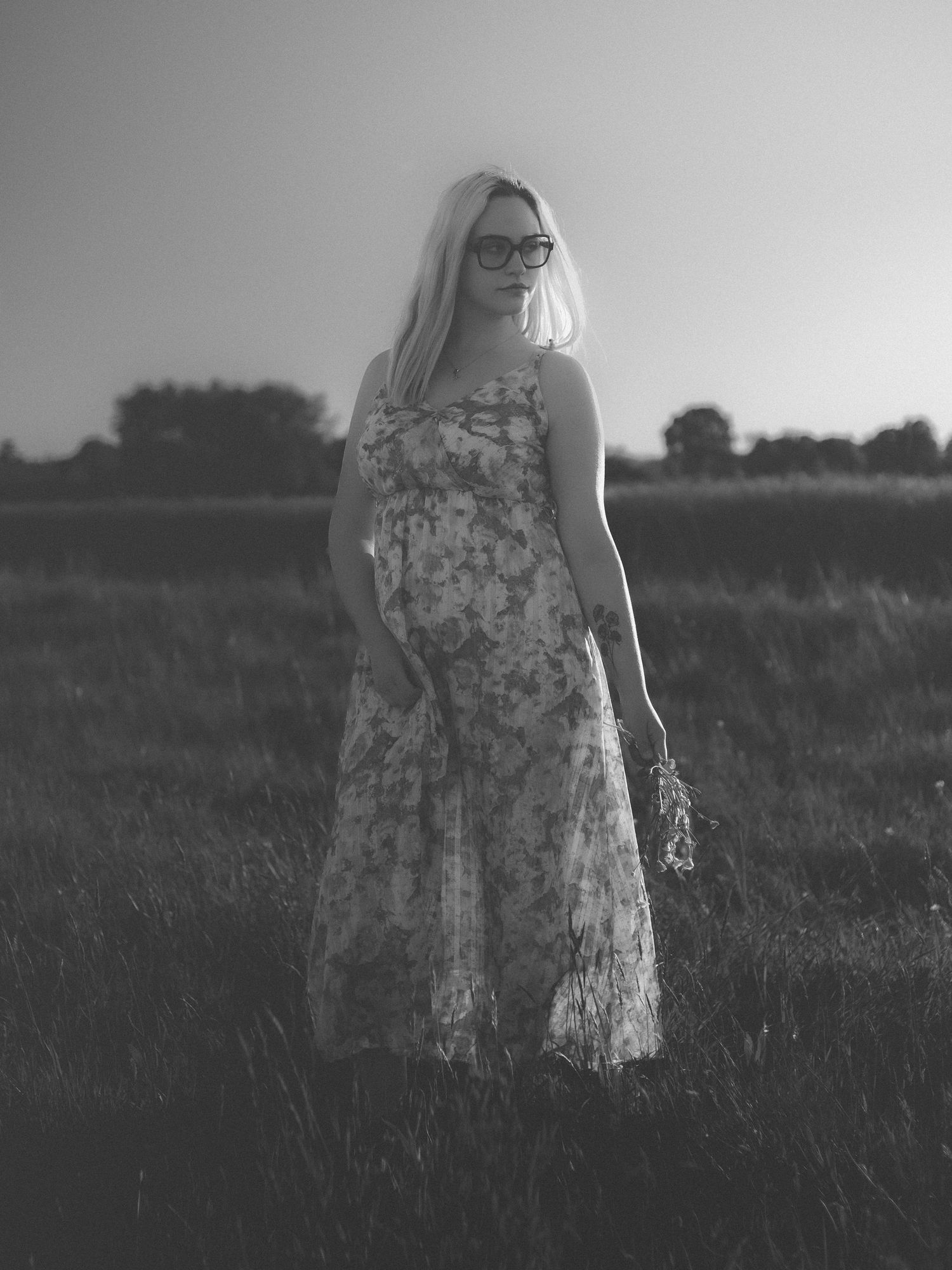 Black and white photo of a blonde woman in a floral dress holding wildflowers in a field at dusk.