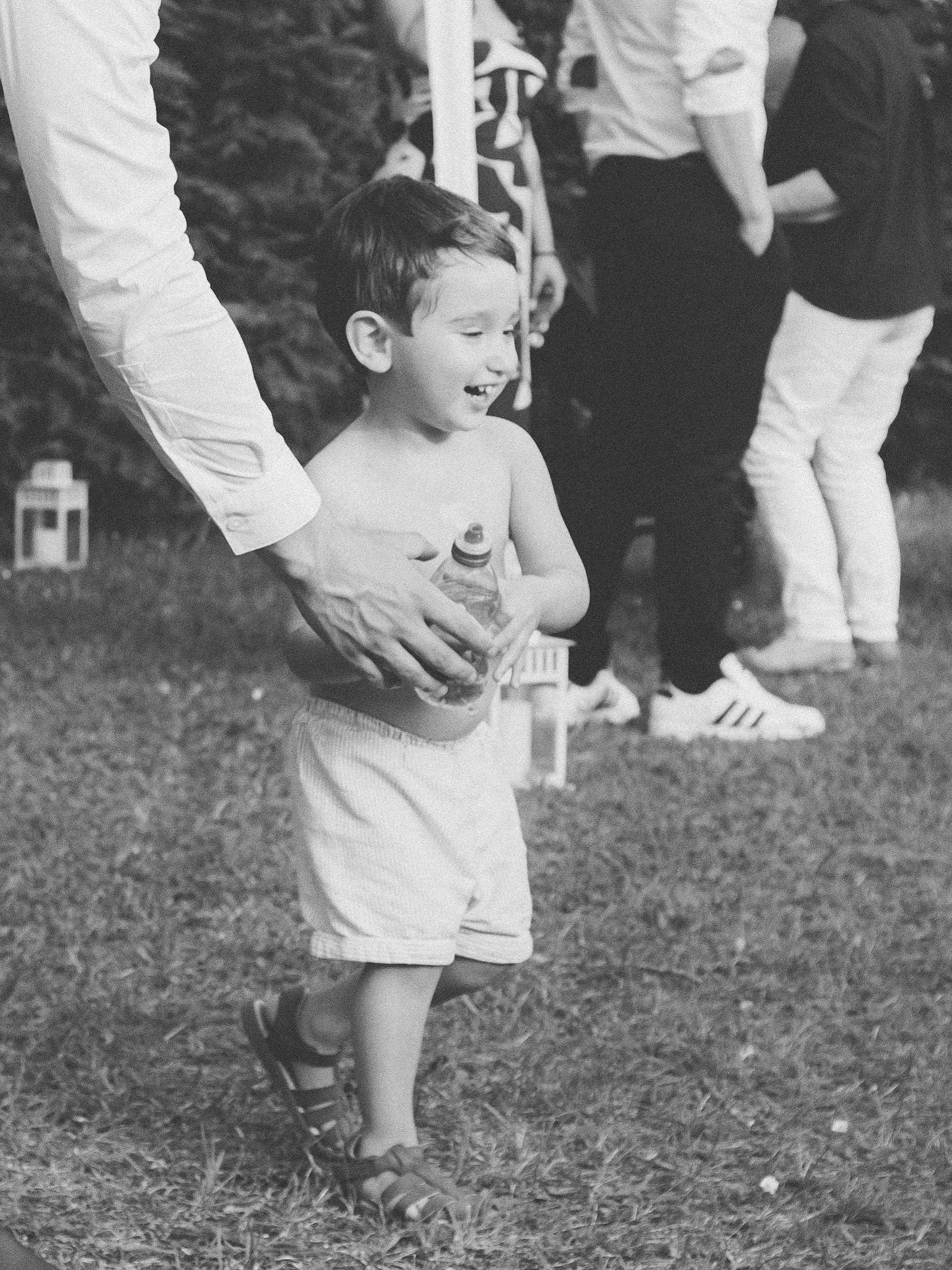 Black and white photo of a shirtless toddler boy crying while holding a bottle at an outdoor event.
