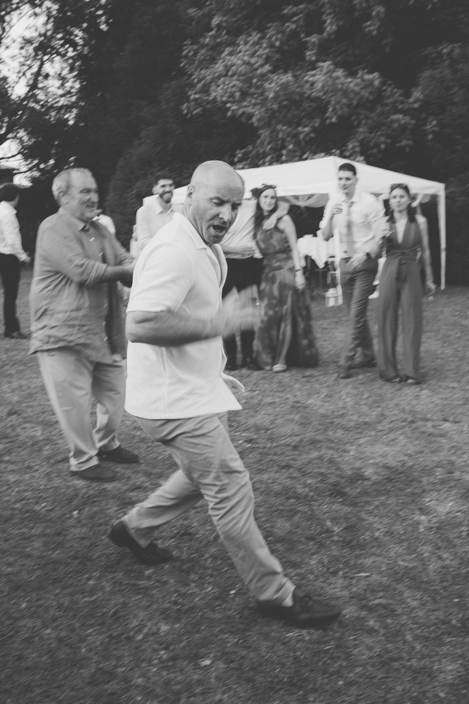 Black and white photo of a bald man dancing energetically outdoors at a garden party with guests watching nearby.