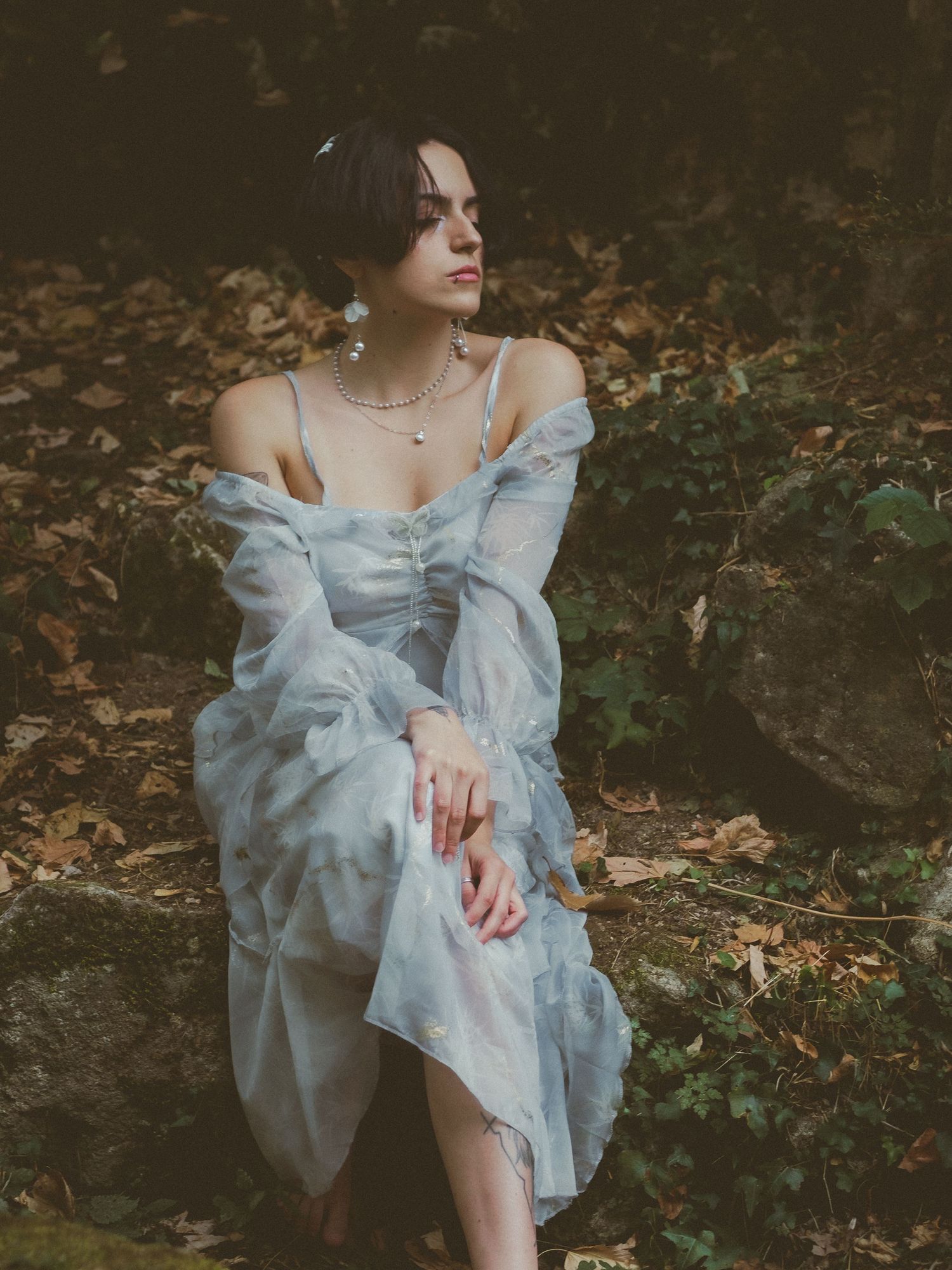 Young woman in blue off-shoulder organza gown sits on mossy rocks surrounded by autumn foliage.