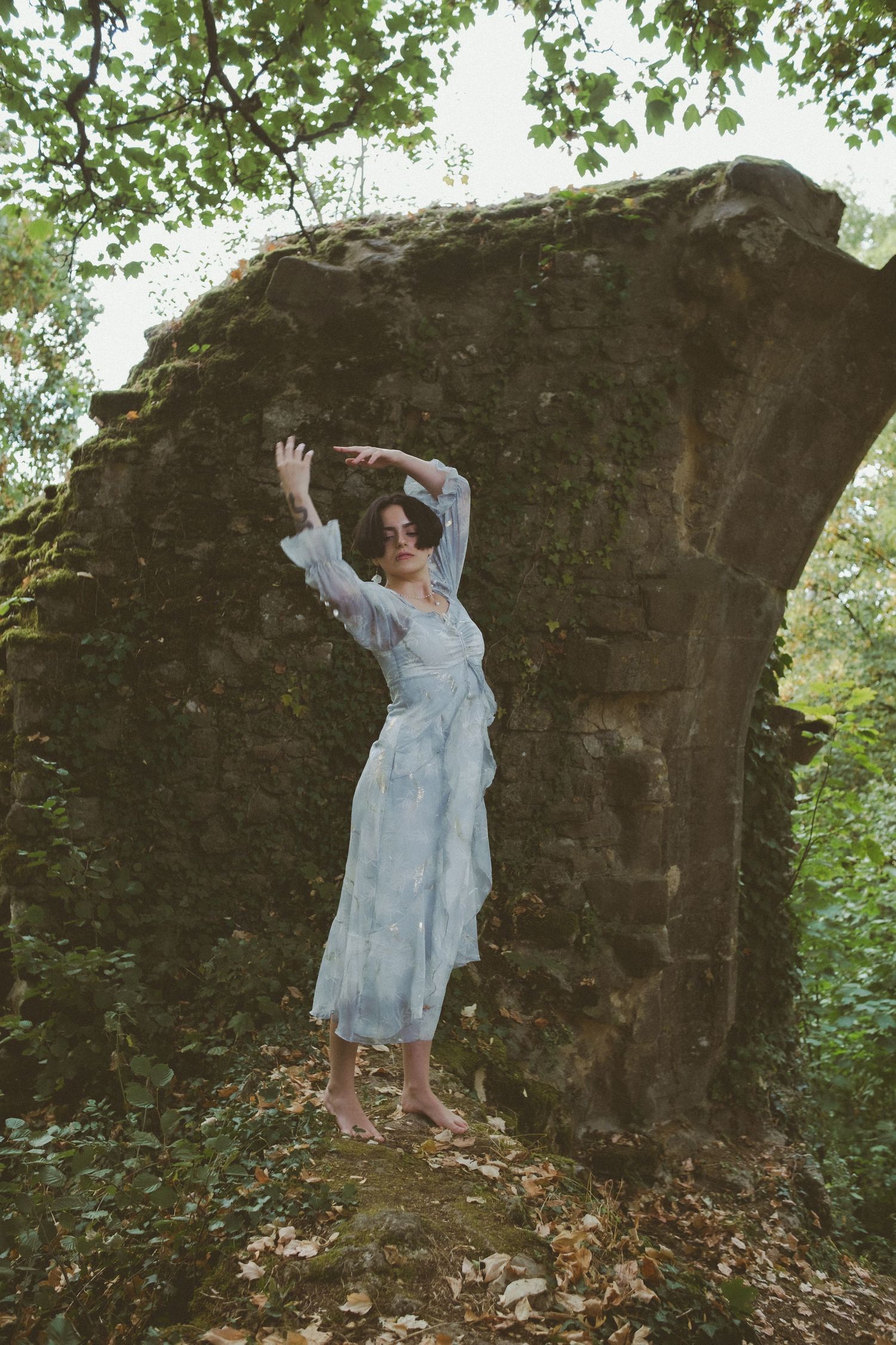 Woman in white lace dress poses barefoot among ancient mossy stone ruins surrounded by lush green forest foliage.