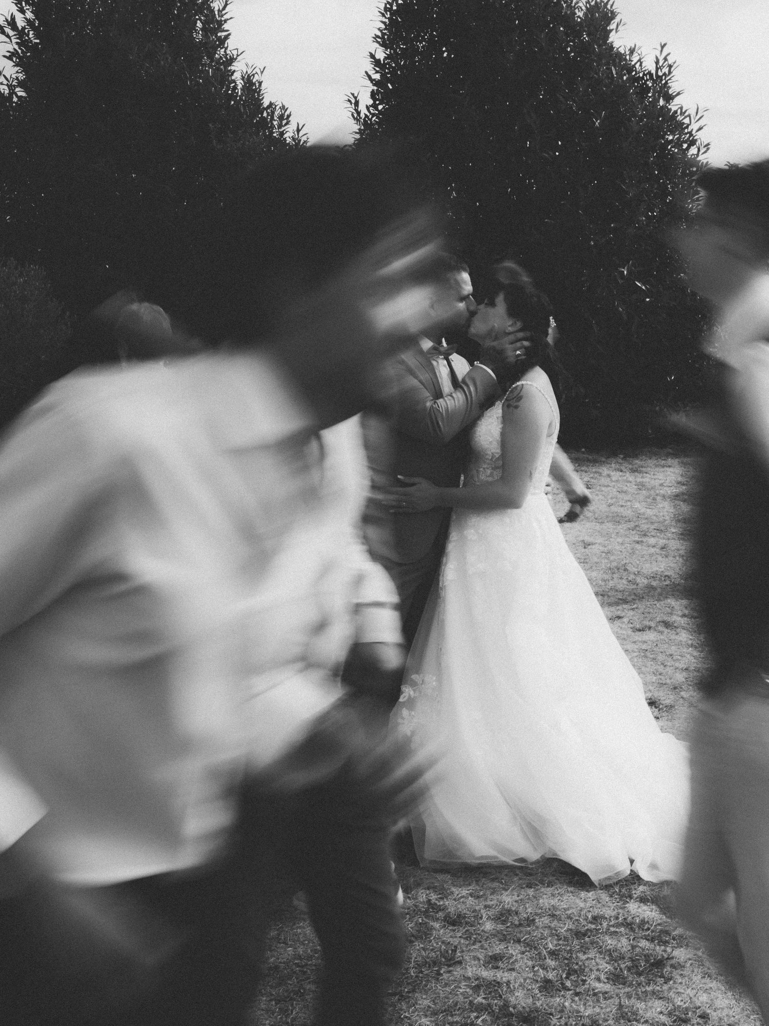 Black and white photo of a bride and groom kissing outdoors, with blurred wedding guests in the foreground.