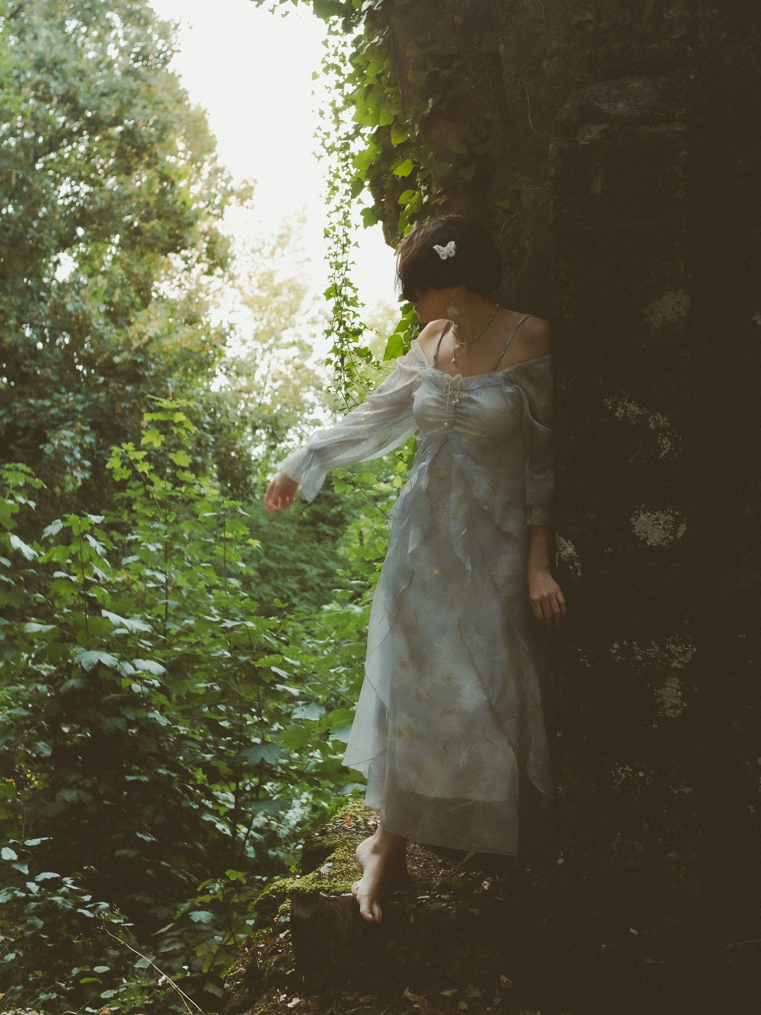 A barefoot woman in a flowing white floral dress stands among lush green forest foliage near a mossy stone wall.