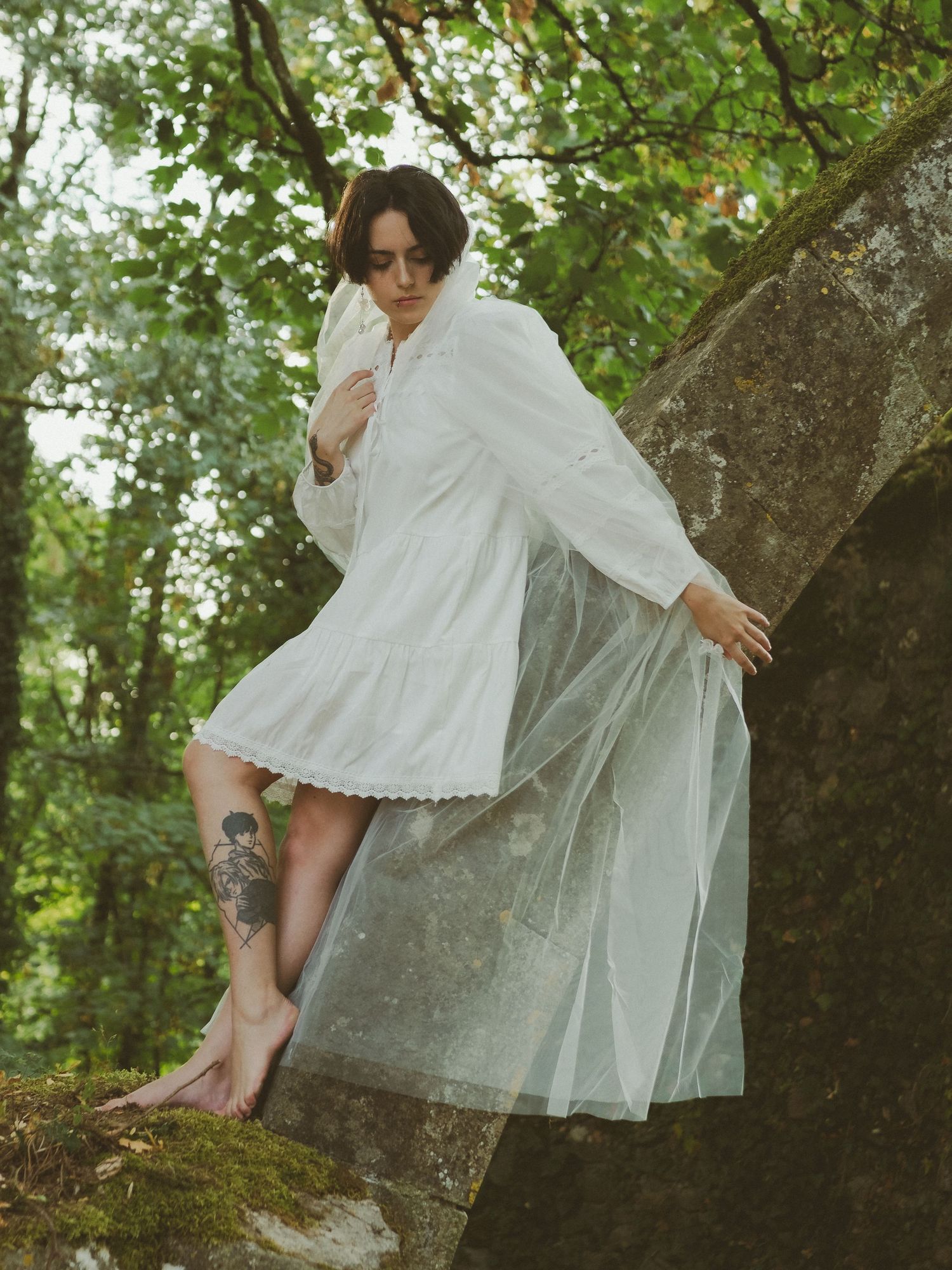Barefoot woman in white dress with sheer overlay climbs mossy rock in forest.