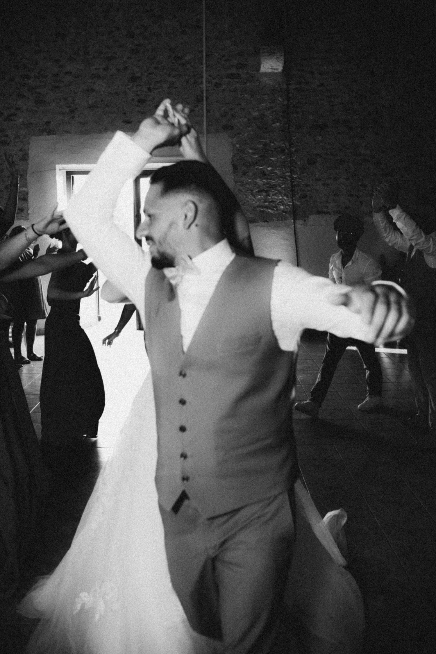 Black and white photo of a groom dancing joyfully with bride at wedding reception, arms outstretched, guests in background.