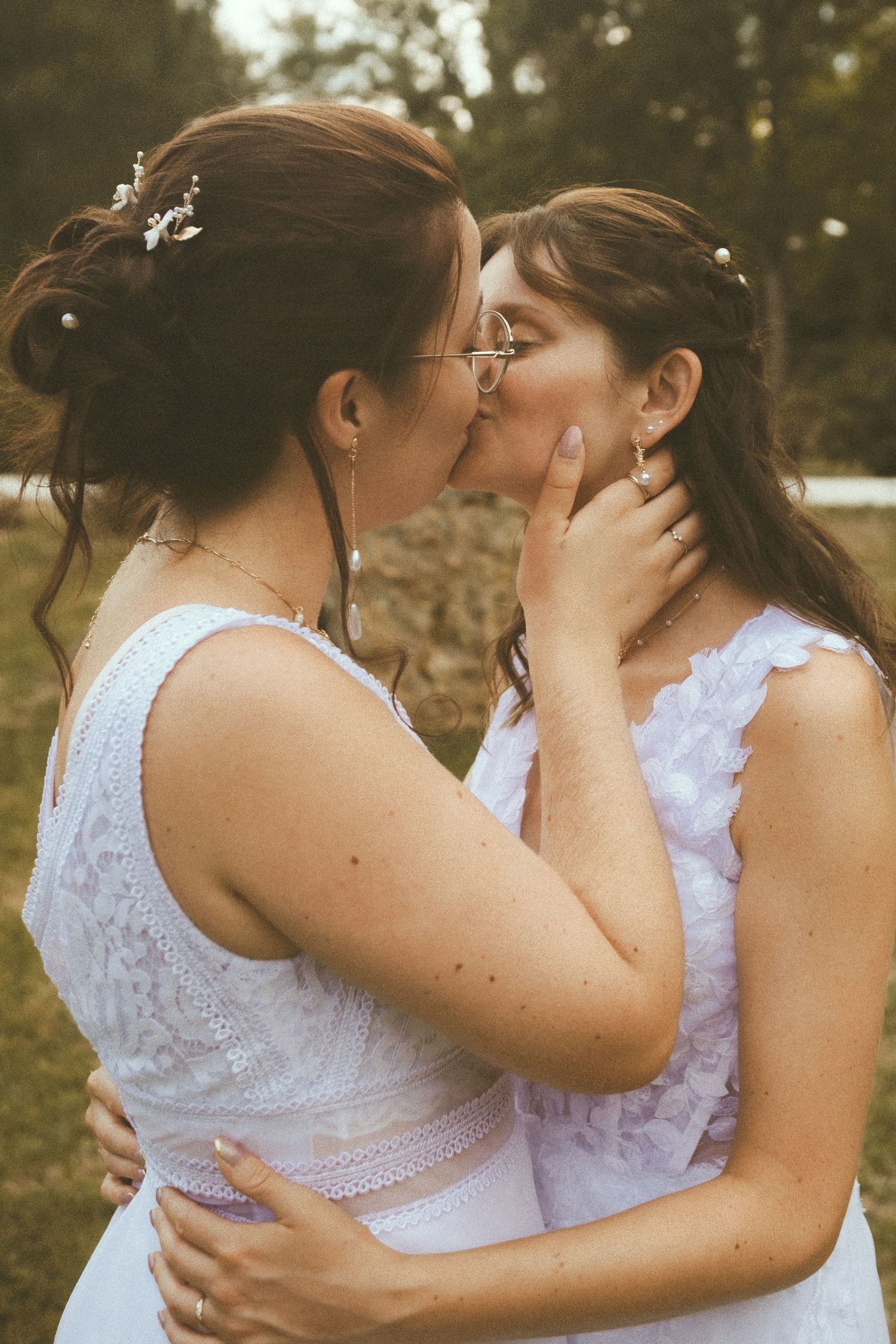 Two brides in white dresses share a romantic kiss outdoors on their wedding day.