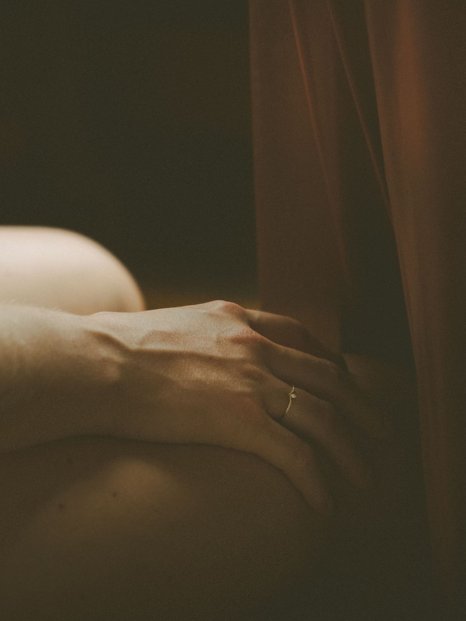 A hand with a delicate ring gently touches warm fabric in soft, moody lighting.