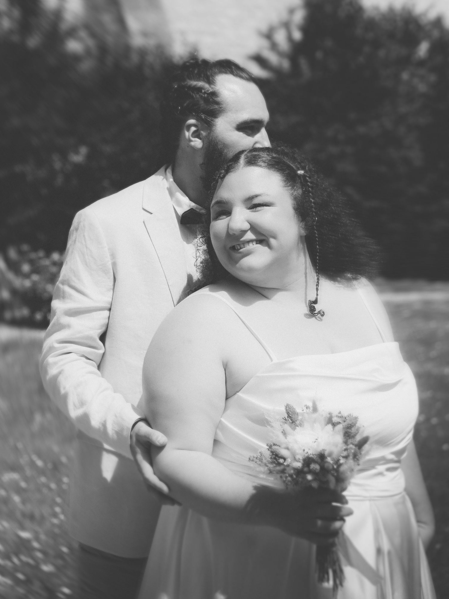 Black and white wedding photo of a smiling bride holding flowers with groom kissing her head.